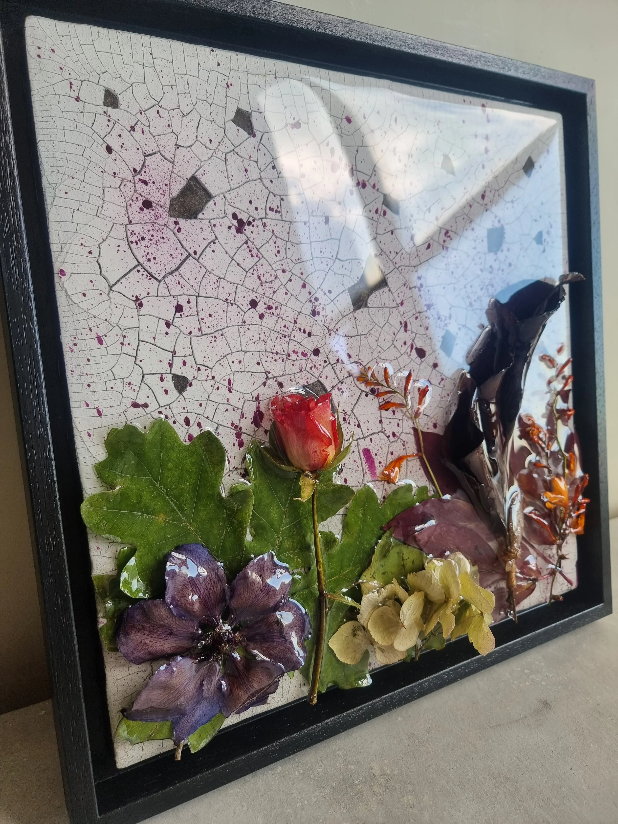A decorative shadow box with a glass front showcasing dried and pressed flowers and leaves, including a purple flower, green leaves, a pinkish-red rosebud, and an arrangement of yellow and brown flowers, with a cracked and splattered background.
