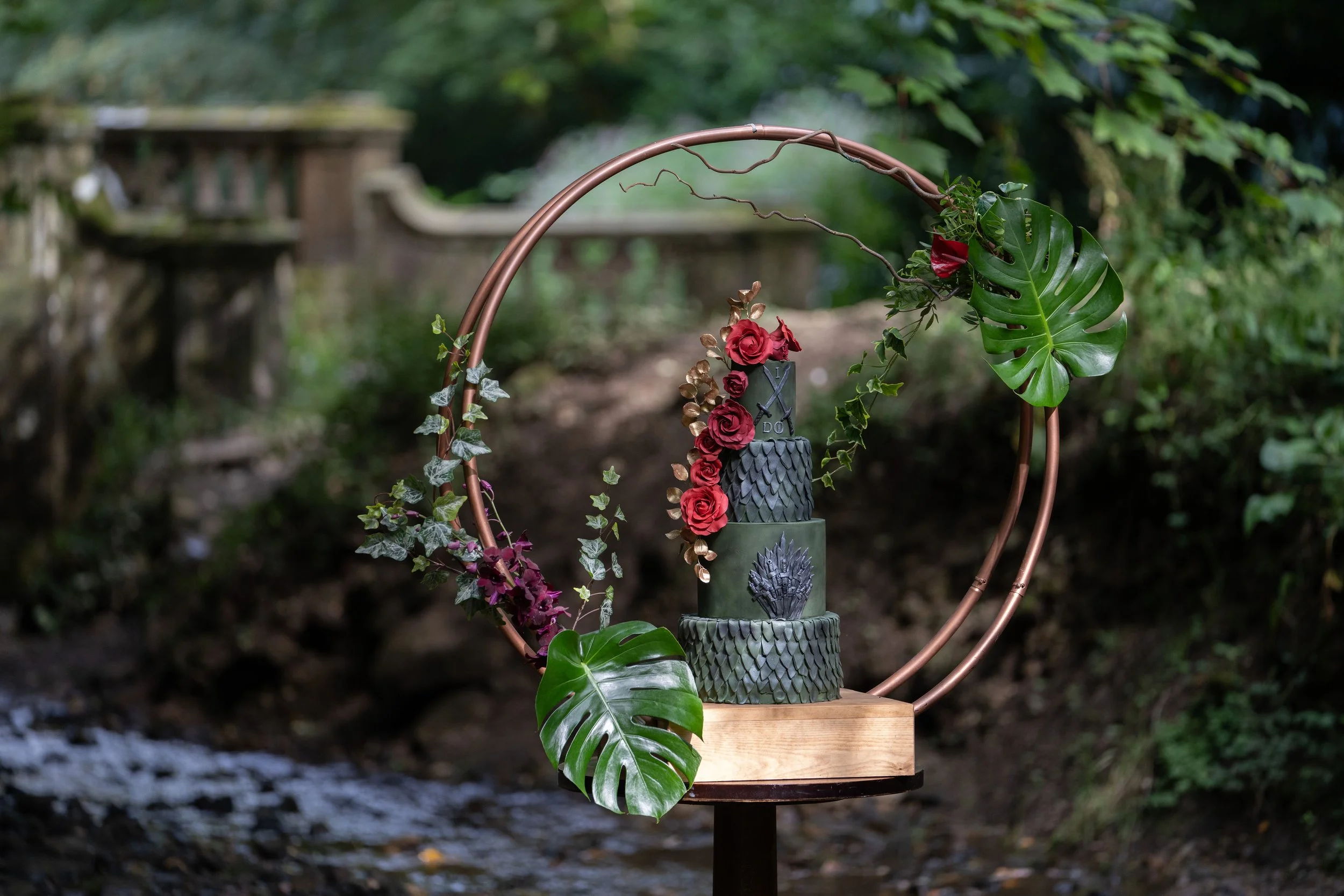 Decorative three-tiered cake with black and gray scales, adorned with red roses and gold accents, set on a wooden base with a copper hoop decorated with large tropical leaves and ivy, against a natural outdoor background.