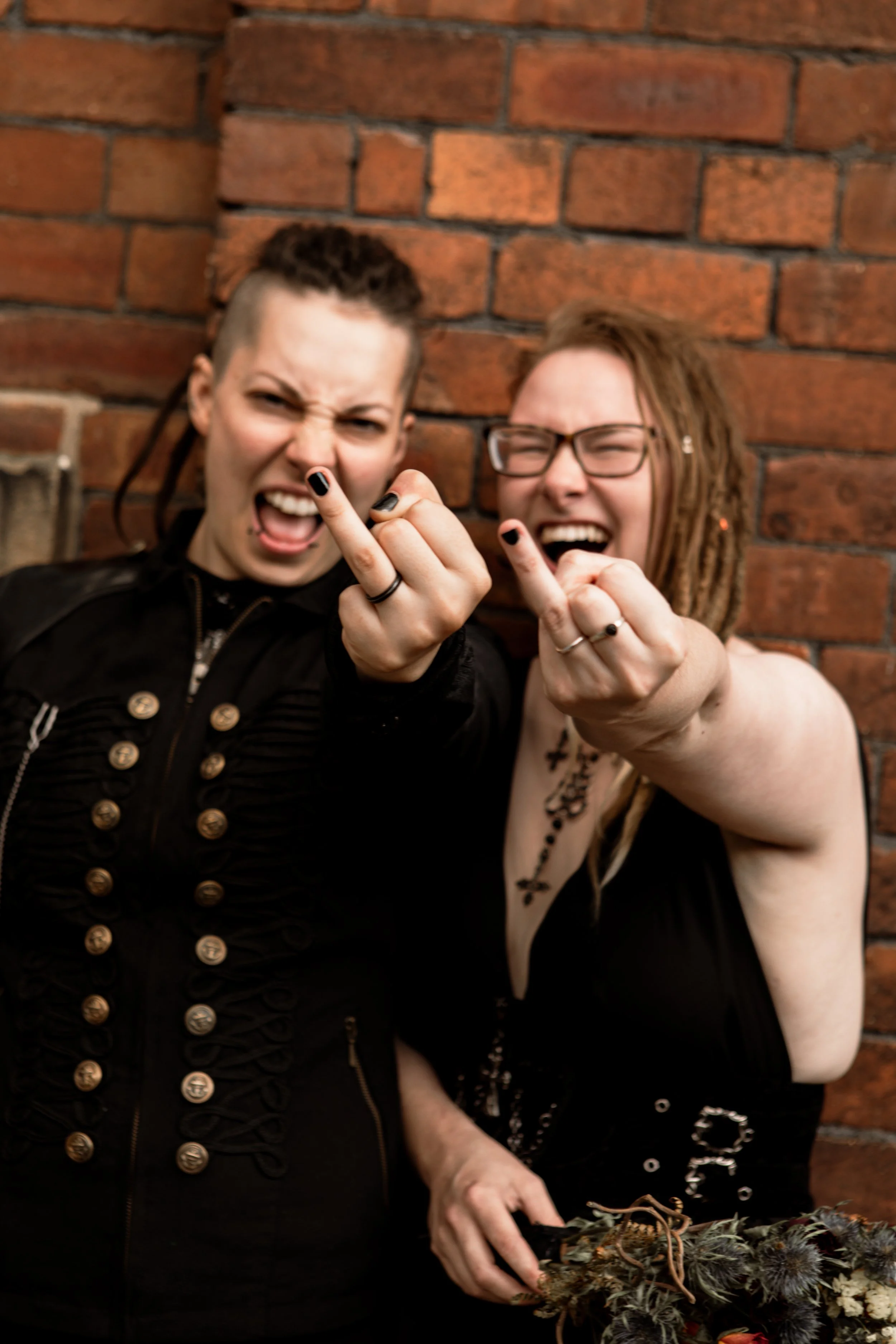 Two women standing in front of a brick wall, both with fierce expressions, raising their middle fingers towards the camera, displaying rings, with dark clothing and makeup.