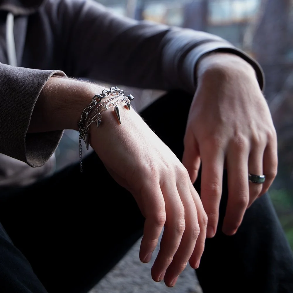 Close-up of a person's hands resting on their lap, adorned with silver bracelets and a ring, with a dark long-sleeve shirt, in front of a window with blurred outdoor background.