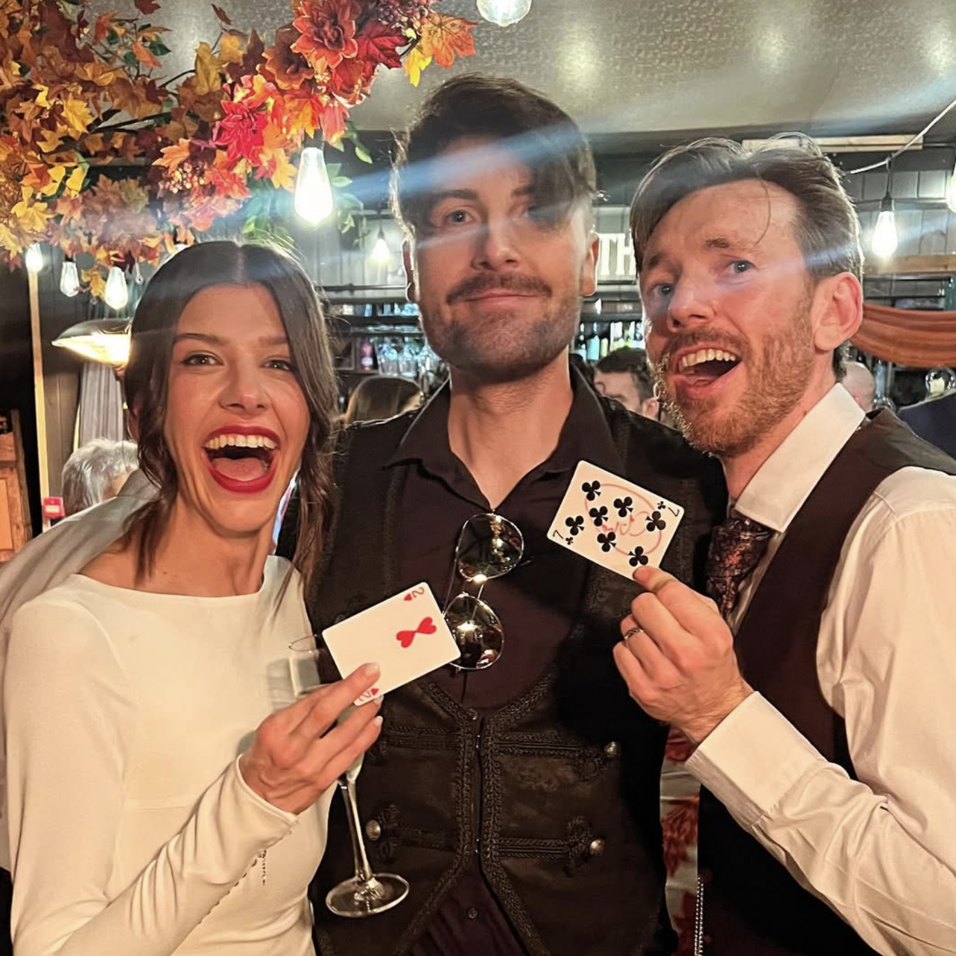 Three people: a woman with dark hair and red lipstick, a man with dark hair and a beard, and another man with silver hair holding playing cards, in a festive setting with autumn decorations and smiling faces.