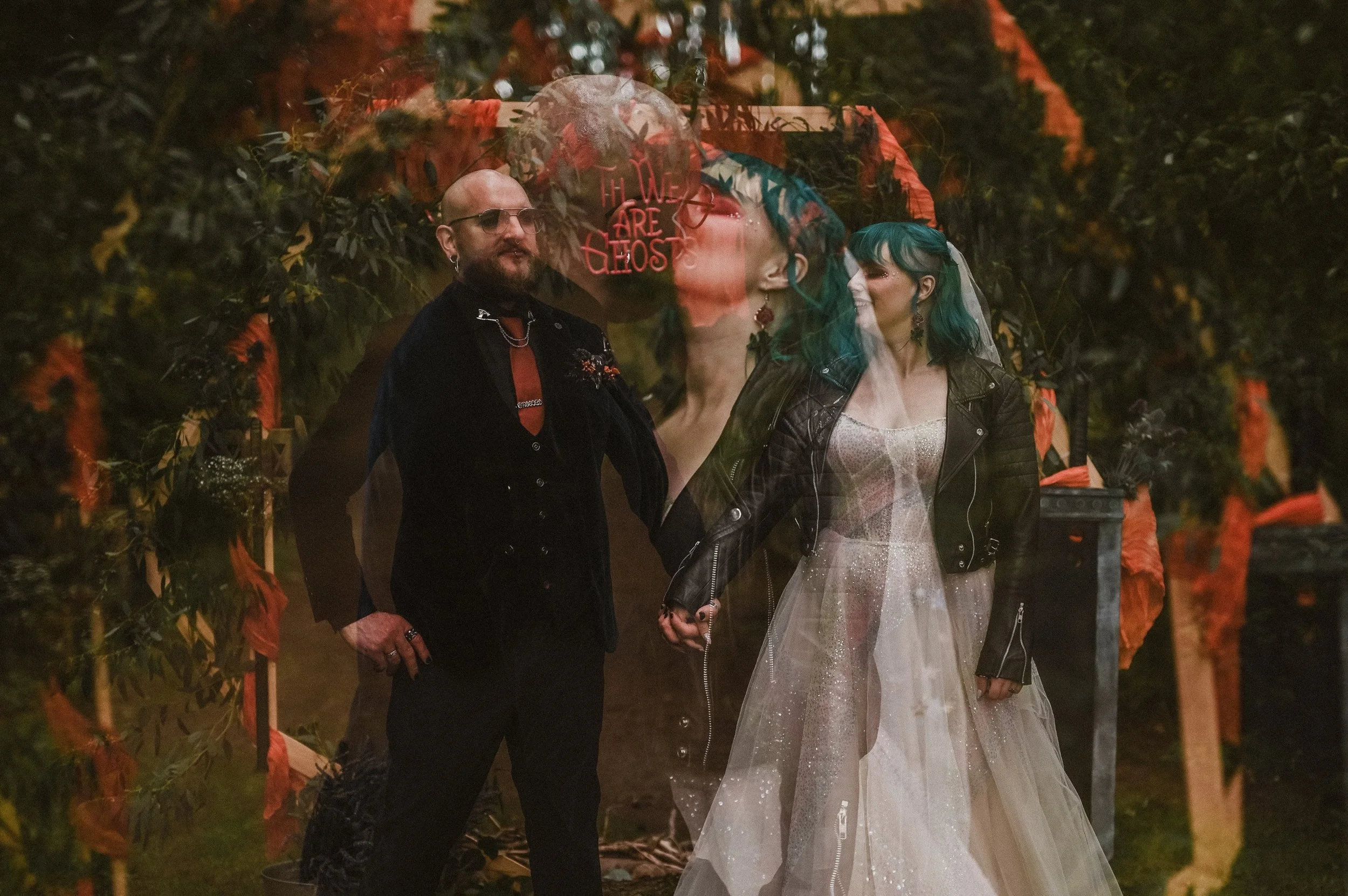 A couple holding hands, dressed in alternative wedding attire, standing outdoors at dusk, surrounded by Halloween or gothic themed decorations and orange ribbons, with a large transparent overlay of a woman with green hair and red earrings, and a neo