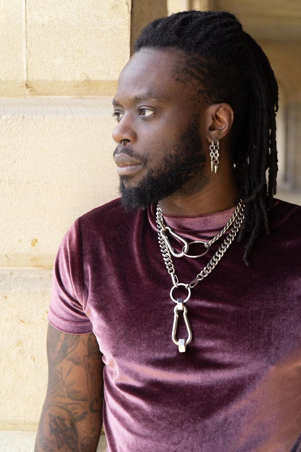 A young man with dreadlocks and facial hair looking to the left, wearing a maroon velvet T-shirt and multiple silver chains and earrings, standing against a beige stone wall.