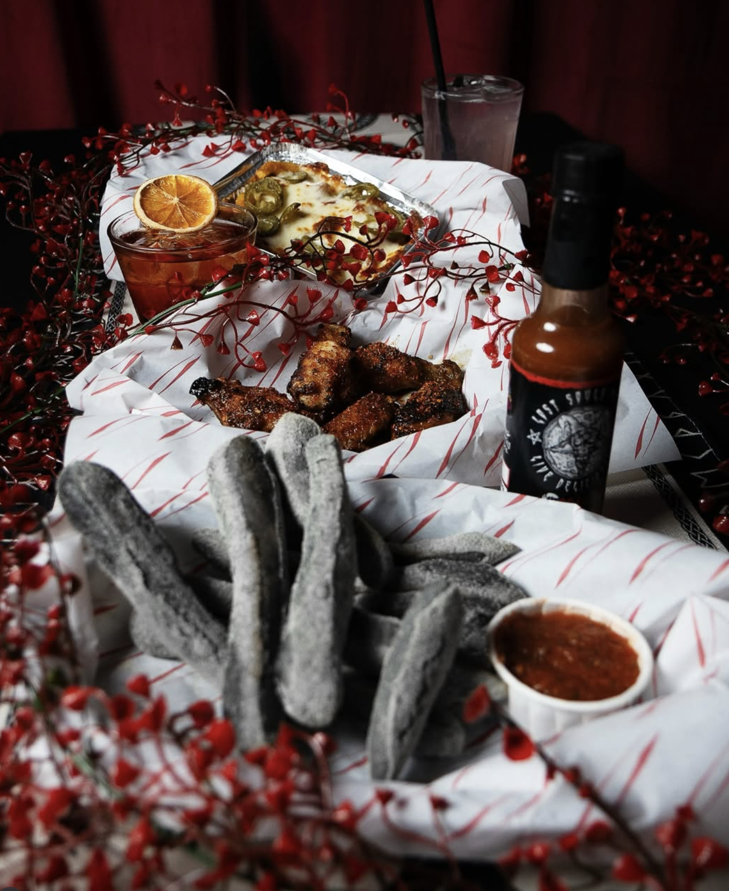 A spread of food on a decorated table including blue corn tortilla chips, spicy chicken wings, dips, beer, and a glass of iced beverage with an orange slice, surrounded by red decorative branches.