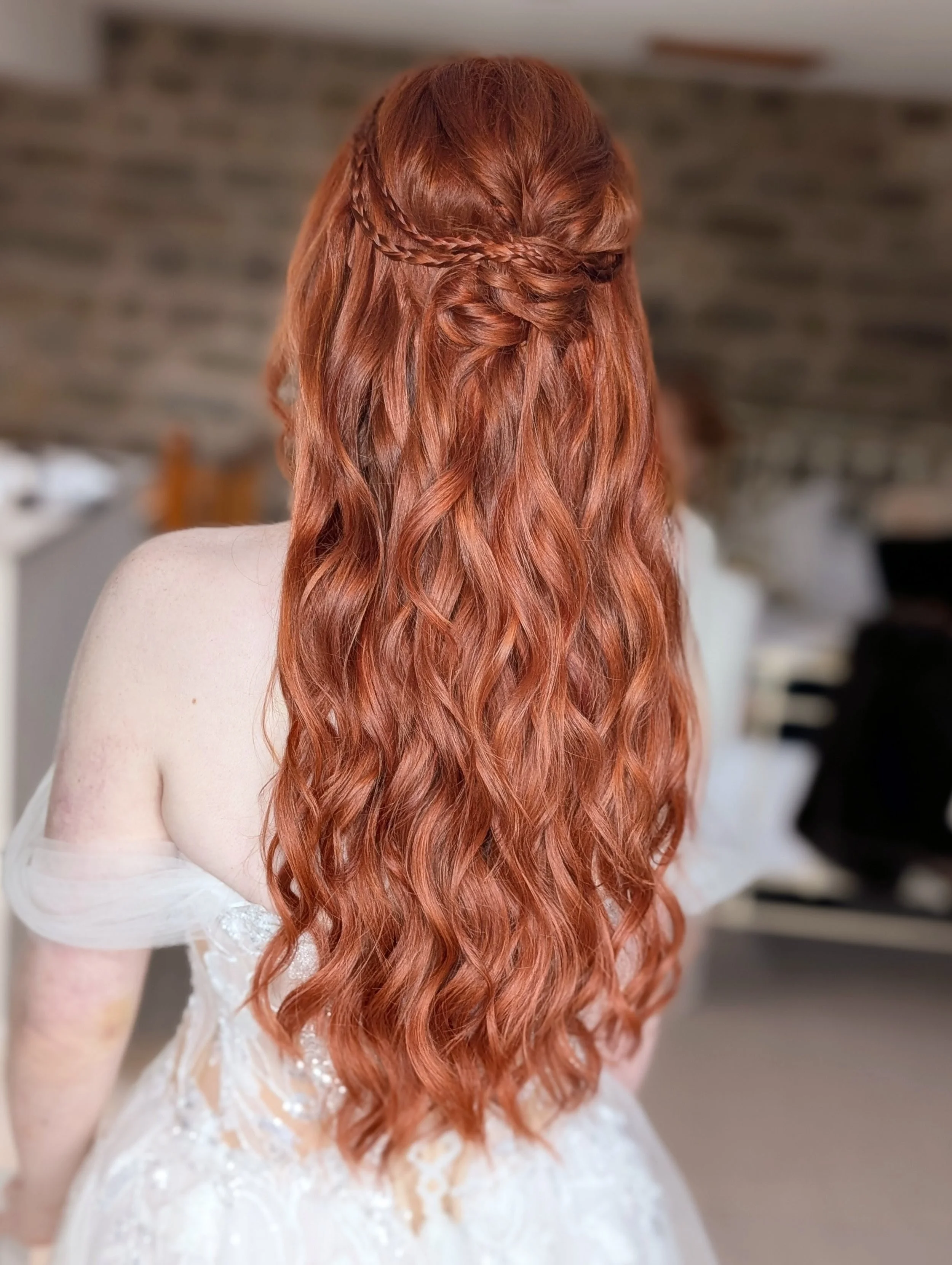 Back view of a woman with long, wavy red hair styled with a braid across the back of her head, wearing a white off-the-shoulder dress, in a room with a blurred background.