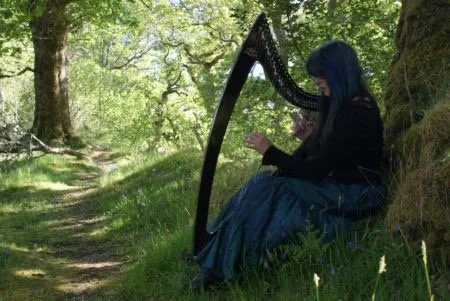 A woman with blue hair playing a harp while sitting on a grassy hill in a forest.