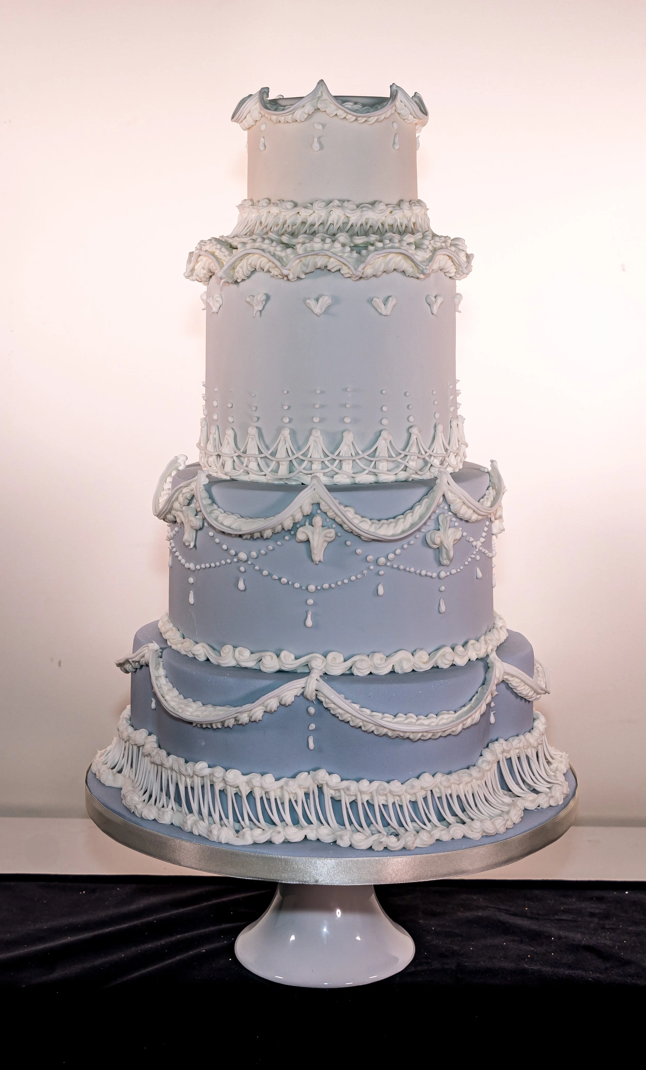 A tall, multi-tiered wedding cake decorated with white icing and intricate piping details, displayed on a white cake stand.