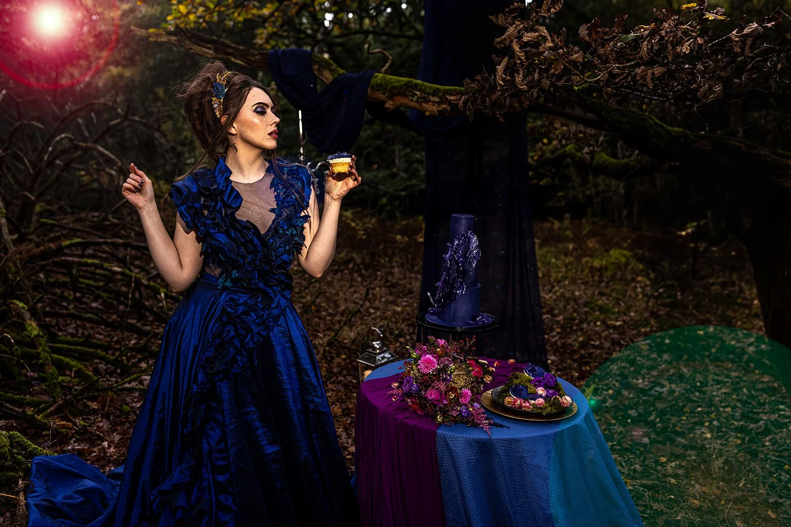 A woman in a dark blue, elaborate gown with ruffled details stands in a forest setting. She holds a slice of cake and looks to her left. A purple and pink floral arrangement is on a table covered with a dark purple and blue cloth, with decorative ite
