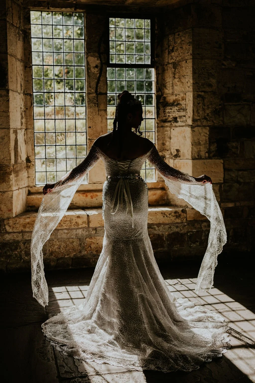 A woman in a wedding dress standing in front of large window with sunlight streaming through, casting shadows on the stone walls and floor.