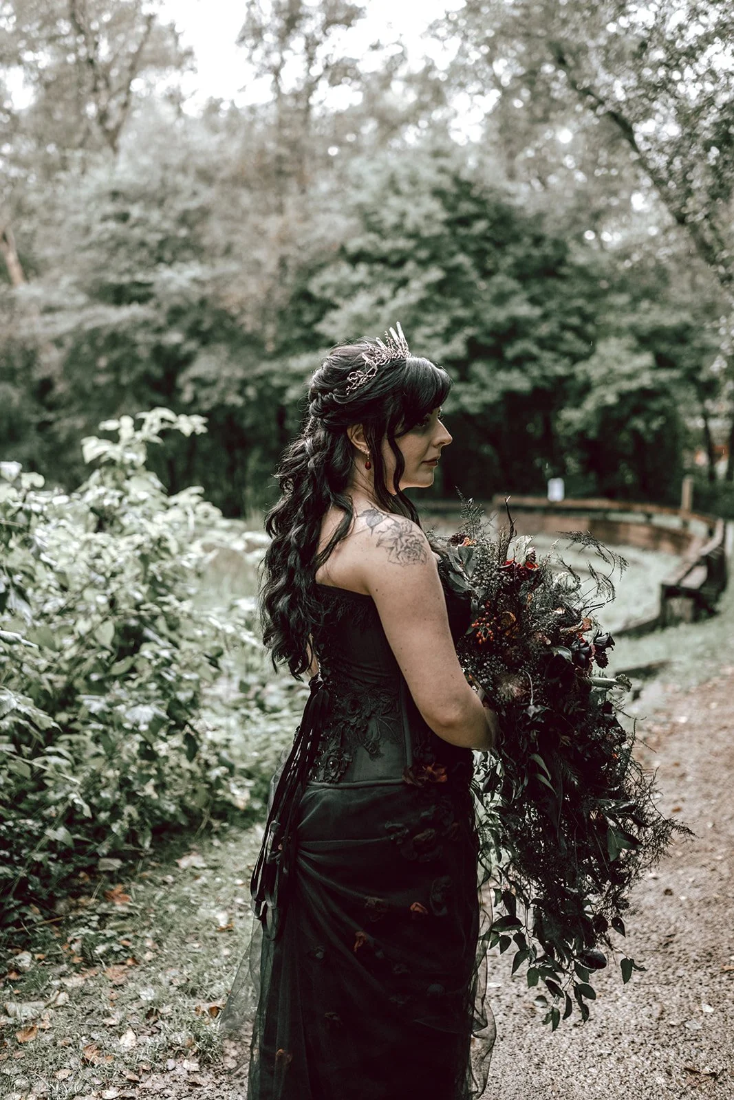 A woman dressed in a black gown, wearing a silver tiara and holding a bouquet of dark flowers and greenery, stands on a wooded path with trees and bushes around her.