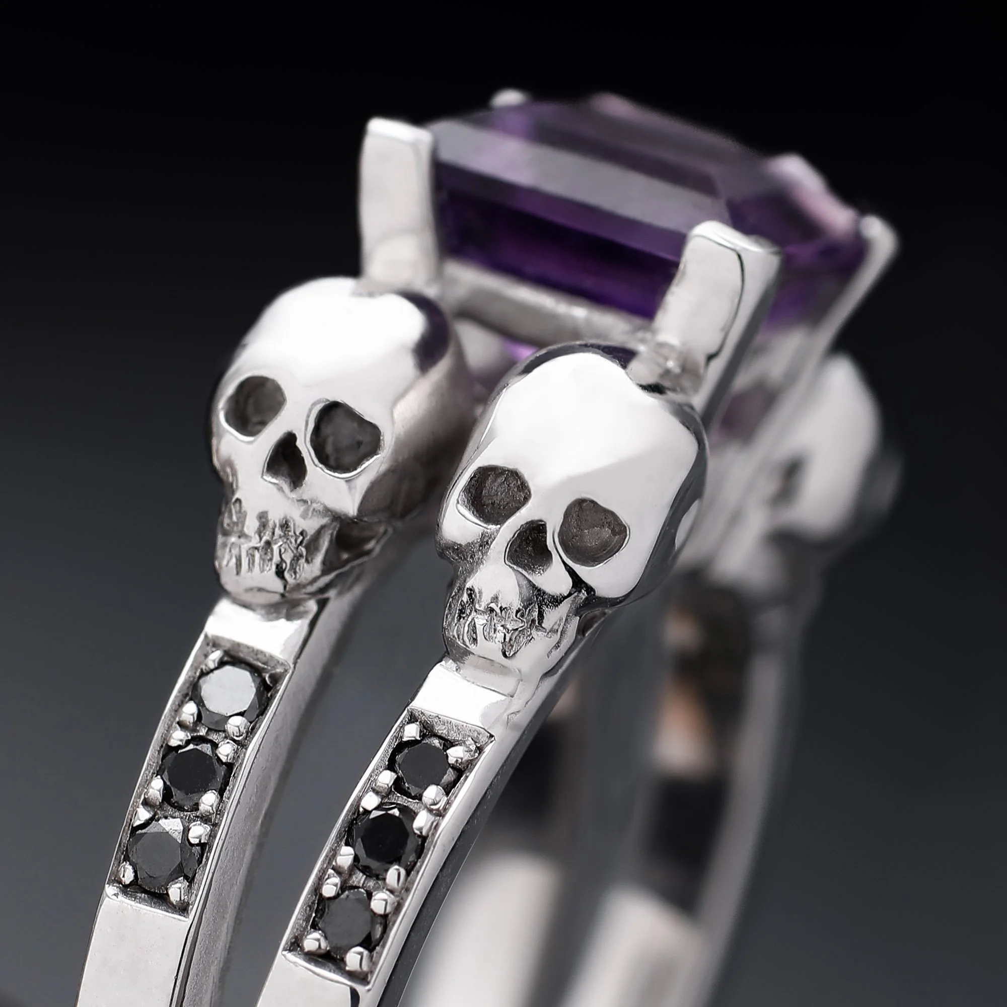 Silver ring with a large purple rectangular gemstone and two skull-shaped accents.