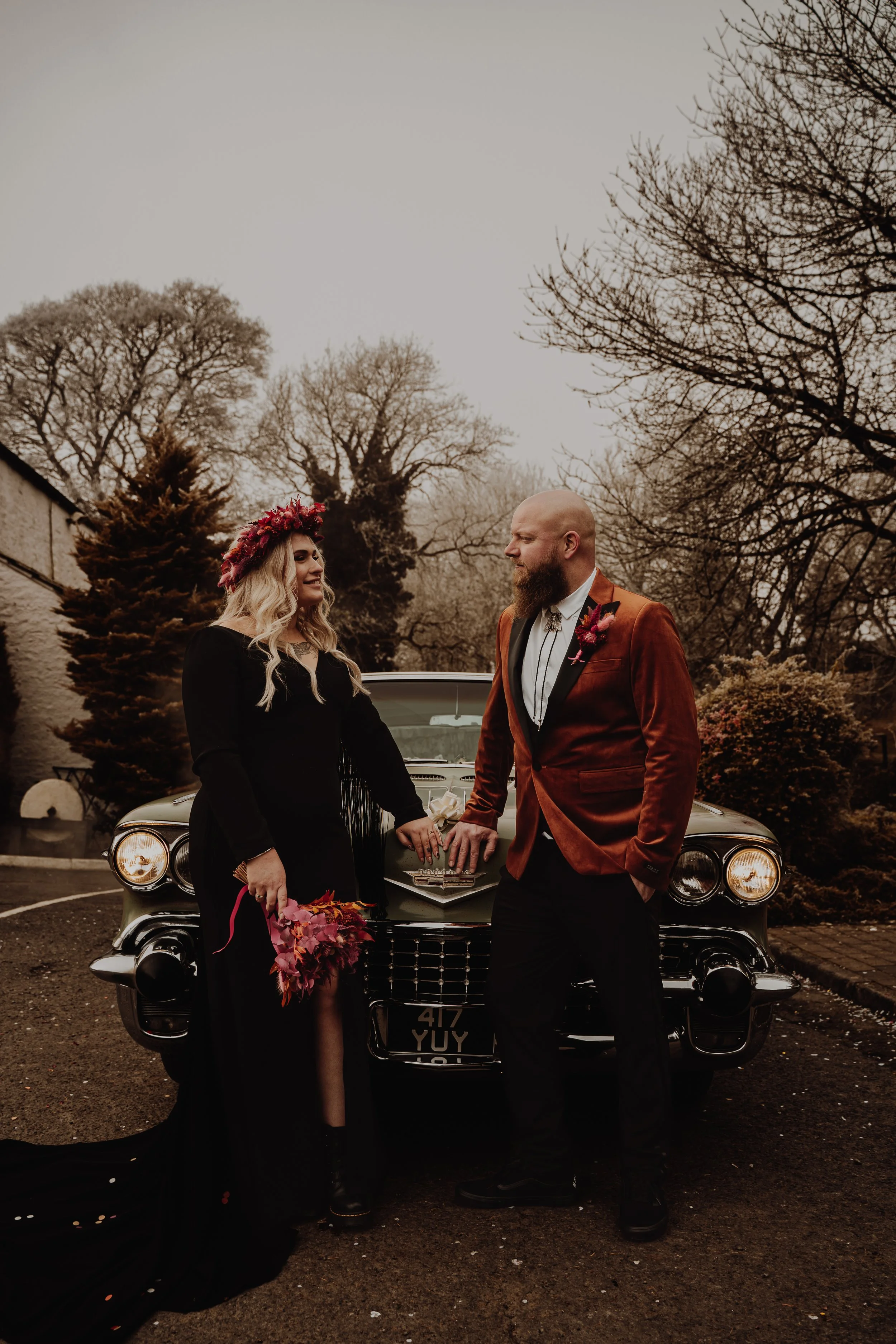 A couple dressed in formal attire standing in front of a vintage car, holding hands, with an outdoor setting featuring trees in the background.