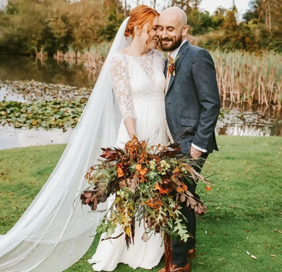 Bride and groom sharing a tender moment outdoors, holding a large fall-themed bouquet of orange, green, and brown foliage, with a pond and trees in the background.