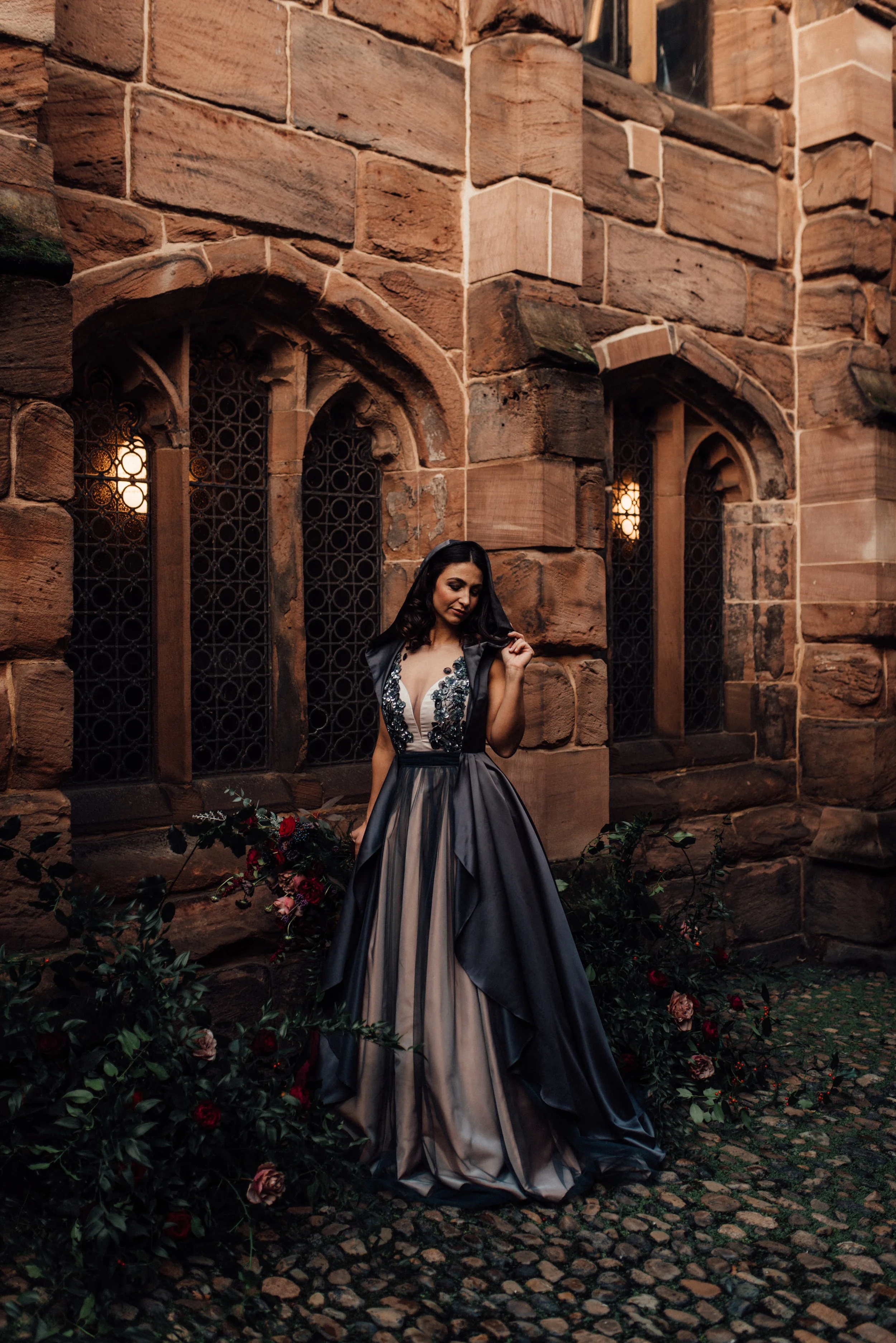 A woman in a black and beige gown standing in front of a historic brick building with gothic windows and surrounded by flowers.