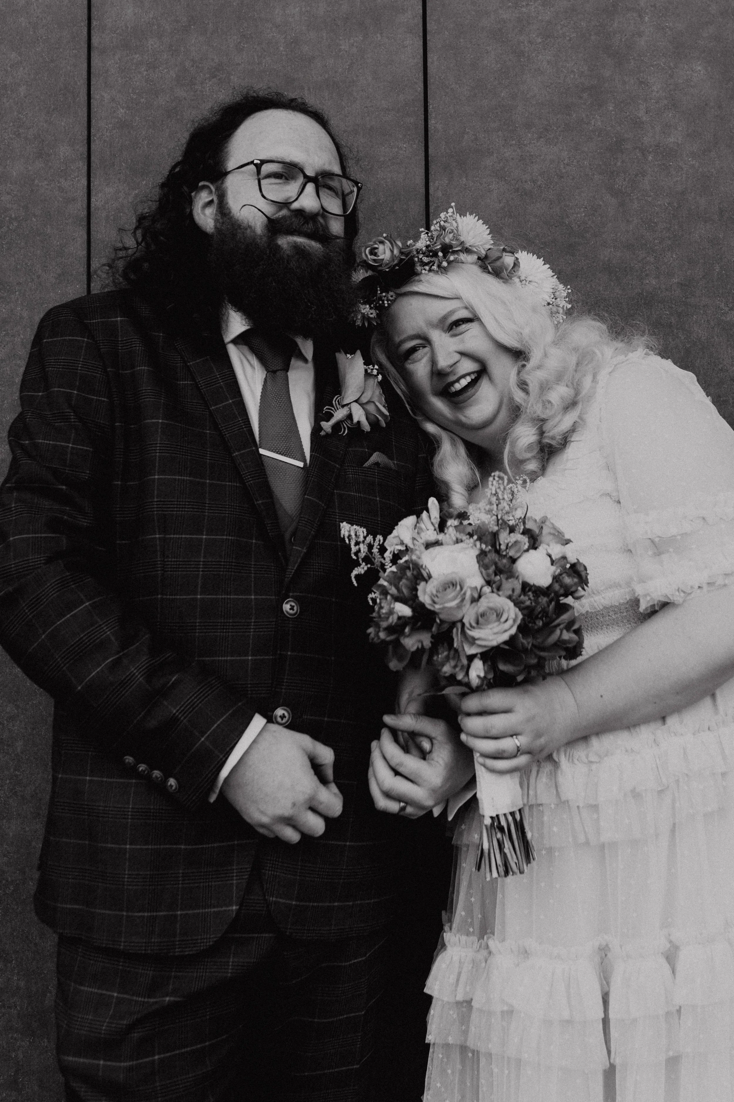 A black and white photo of a joyful couple on their wedding day, with the woman holding a bouquet of flowers and wearing a floral crown, and the man dressed in a checked suit and glasses, both smiling.