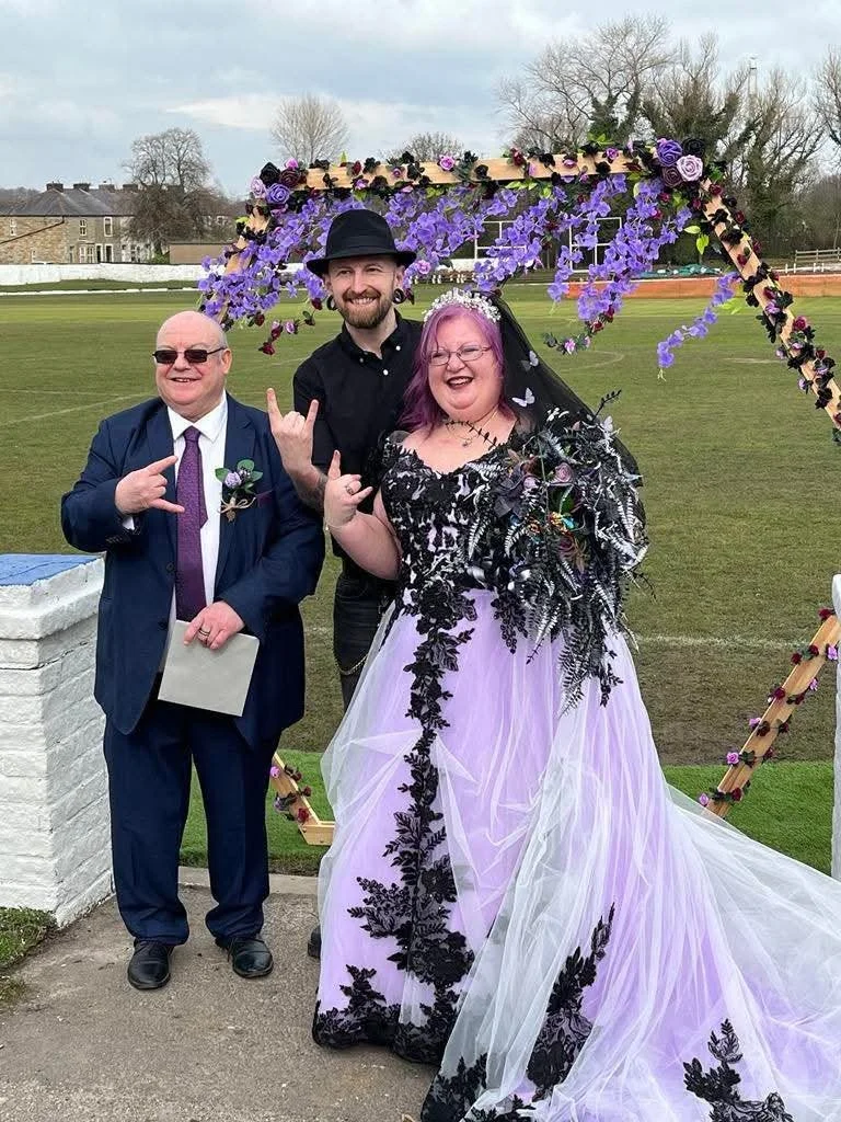Three people at a wedding celebration posing in front of a floral arch; the bride wears a purple and black dress with black lace details, the groom wears a suit and tie, and another man in the middle wears a black shirt and hat; all are smiling and m