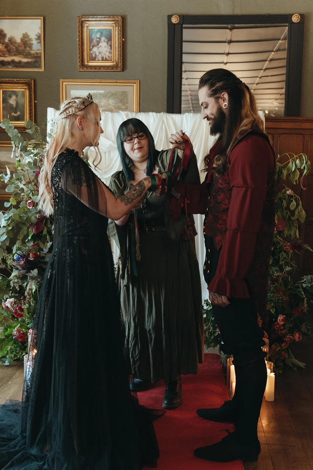 Three people at a wedding ceremony exchanging rings. The bride has long blonde hair, a crown, and a black dress. The groom has long hair, a beard, and a red and black outfit. The officiant, in the middle, has dark hair with streaks and glasses, and i