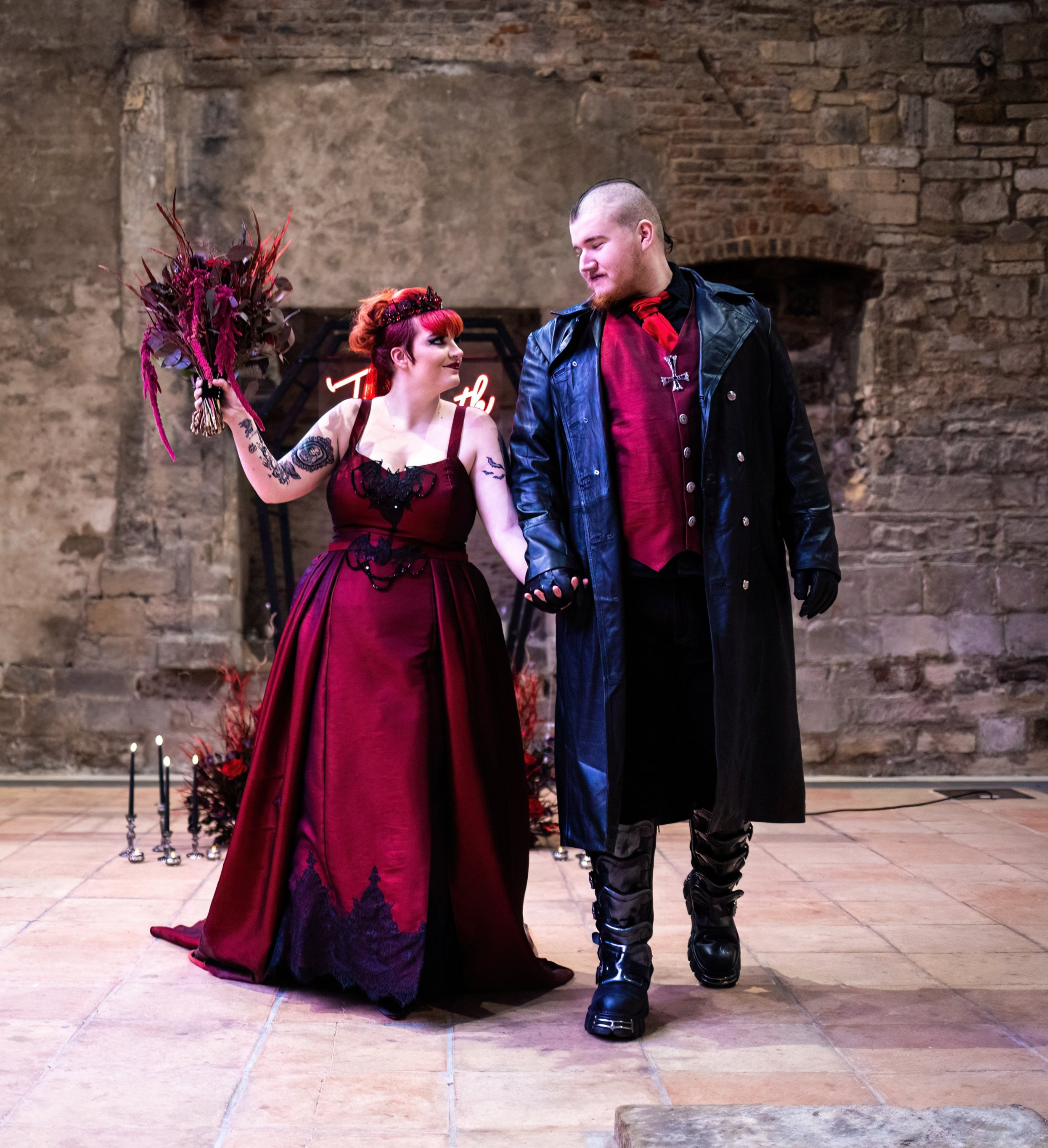 Couple holding hands at a gothic-themed event or performance. The woman in a red dress with black embroidery, holding a bouquet, and the man in a long black leather coat and red shirt, with gothic accessories, standing in front of a brick wall with c