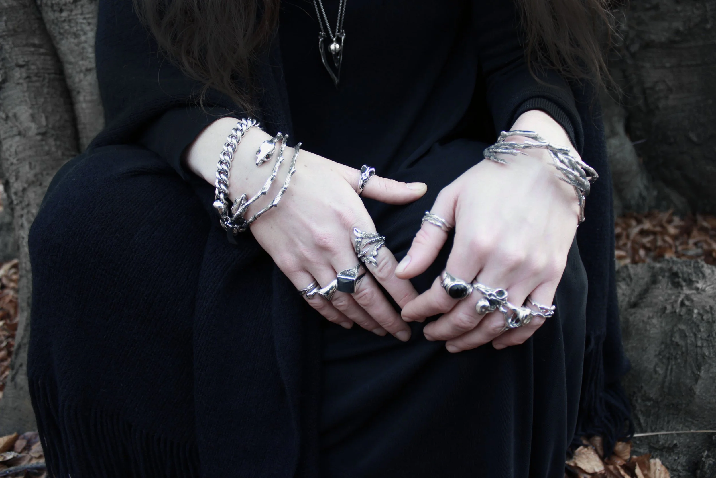 Person wearing multiple silver rings and bracelets, sitting outdoors with a tree trunk and fallen leaves in the background, dressed in black.