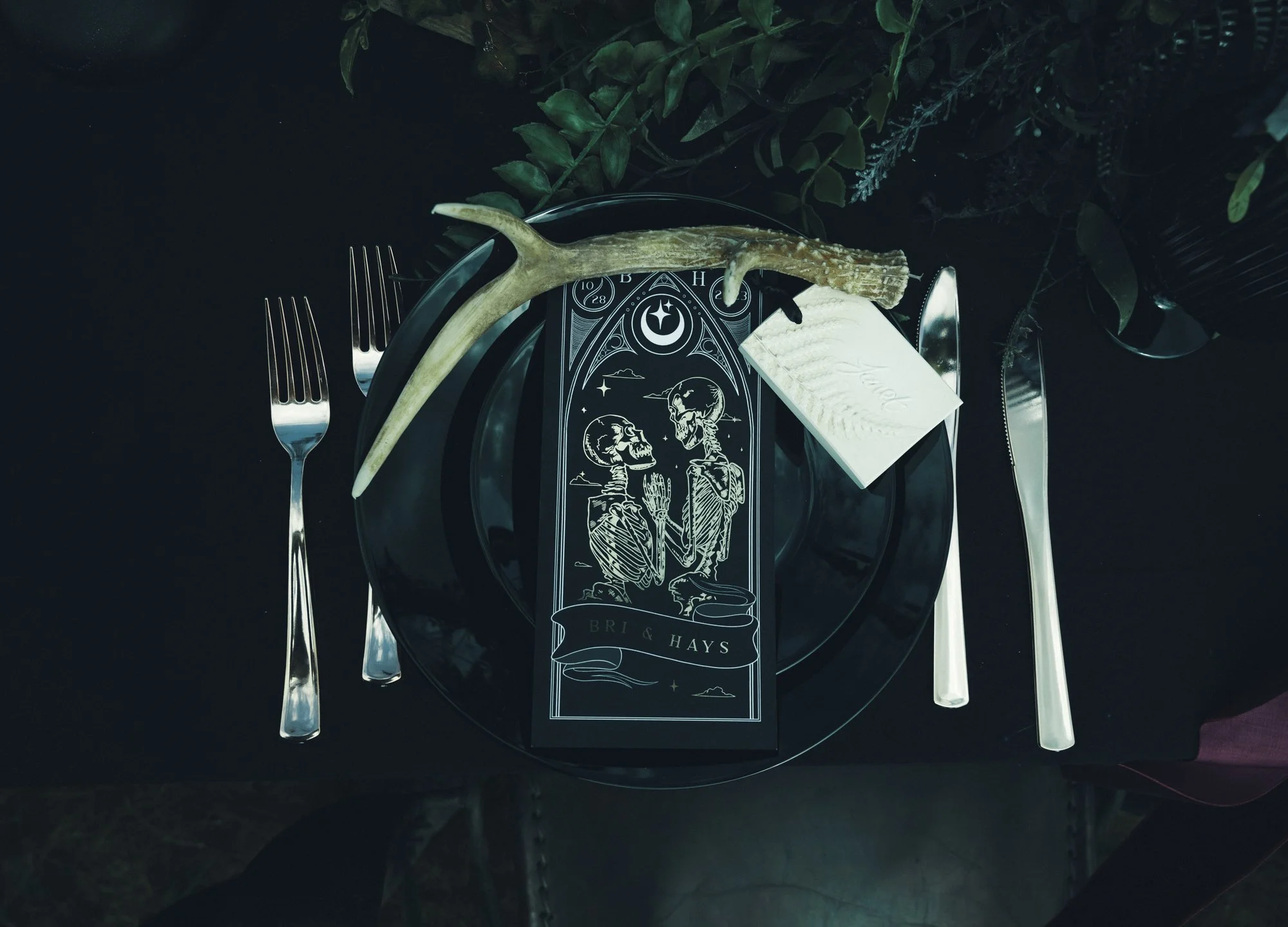 A black table setting with silver forks, knives, and a black plate. A black card featuring a skeleton couple holding hands and the text 'Bri & Hays' is placed on the plate. Additionally, there is an antler on top of the card and a white place card ne