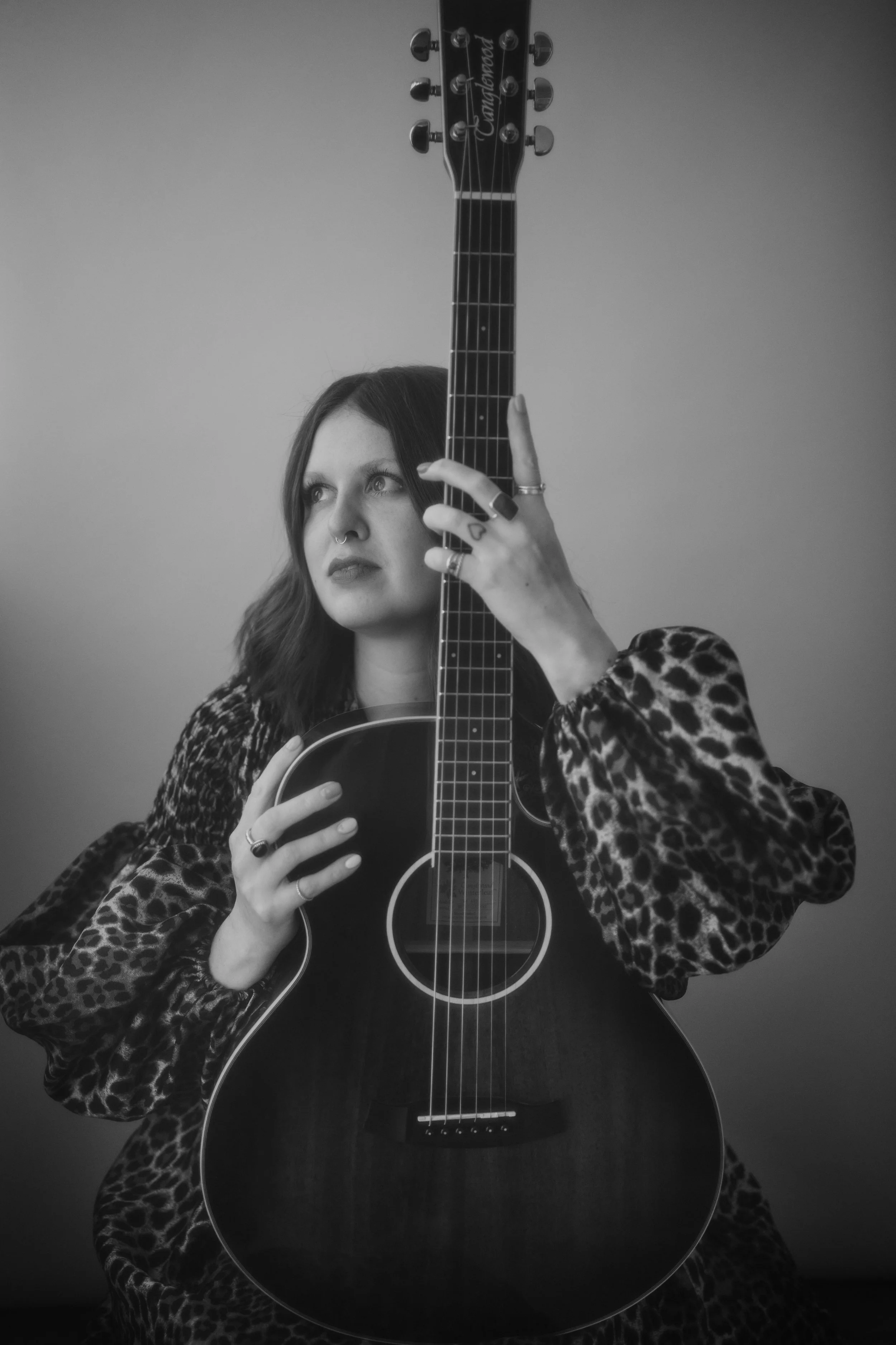 A woman with long hair and a nose piercing, wearing a leopard print jacket, is holding an acoustic guitar in front of her face, with her left hand on the guitar neck and her right hand on the body of the guitar. The image is in black and white.