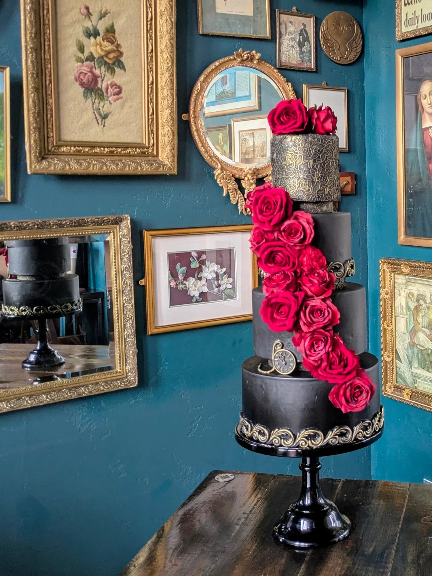 A tall, multi-tiered black cake decorated with pink roses and gold accents on a black cake stand, against a background of framed art and mirrors with ornate gold frames.