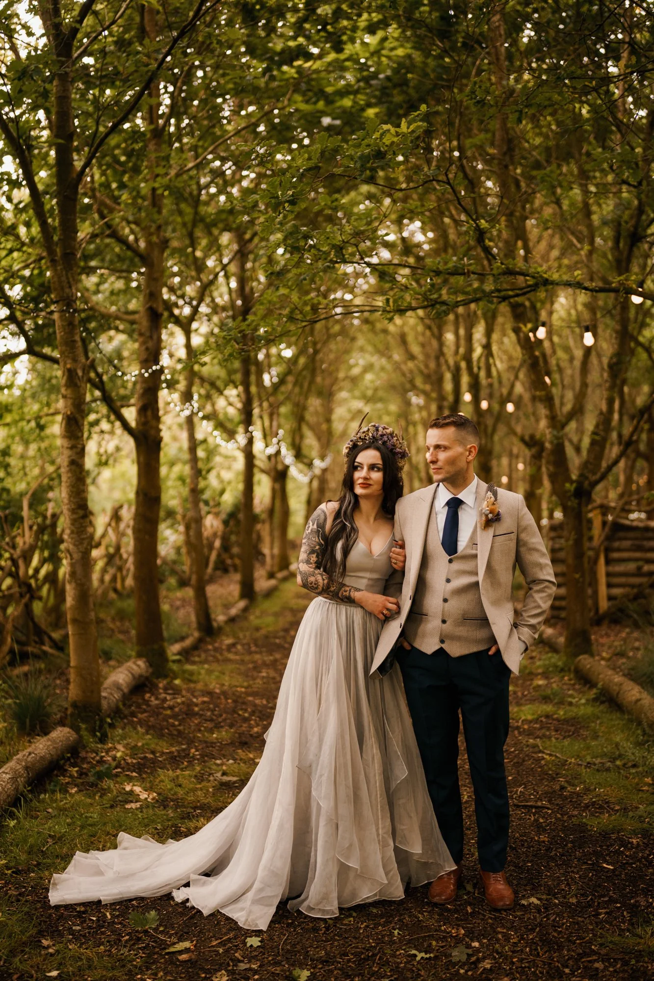 A bride and groom walking arm-in-arm through a wooded pathway decorated with string lights for their wedding.