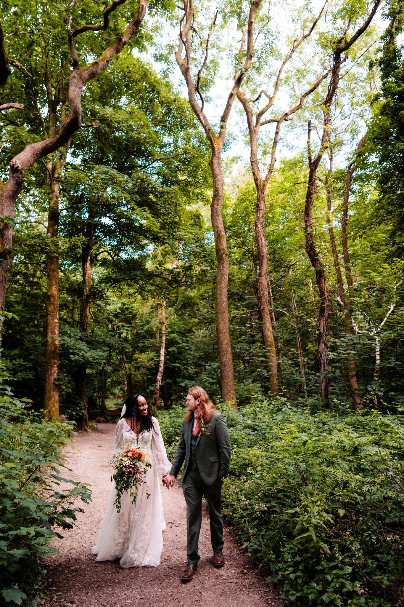 A newly married interracial couple walking hand-in-hand through a lush green forest, the bride in a white lace wedding gown holding a large bouquet, and the groom in a dark suit with a boutonniere.