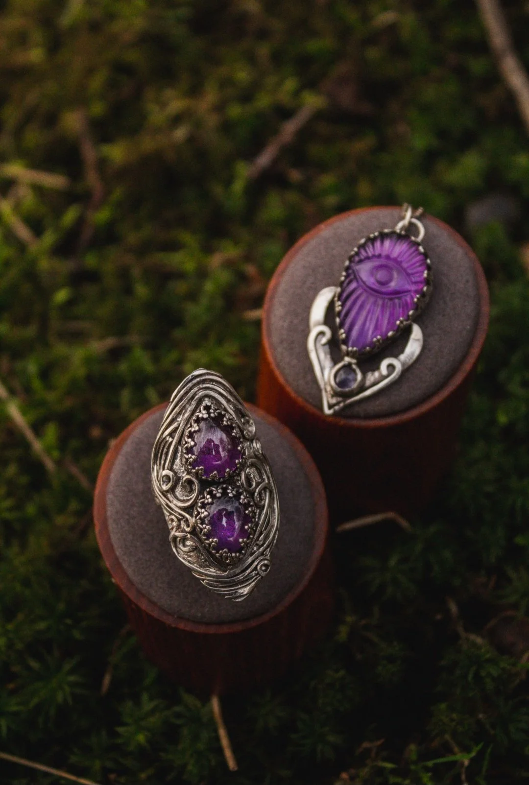 Two silver rings with purple gemstones resting on small wooden stands on green mossy ground