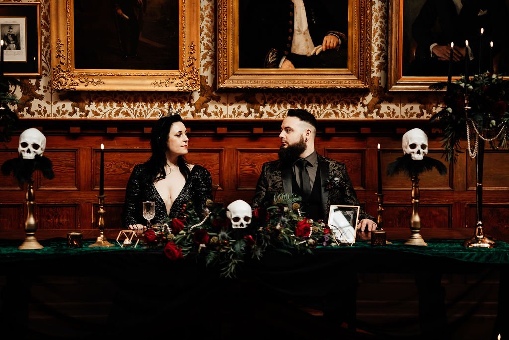 Gothic couple sit at the top table at their wedding gazing at each other. Their table is adorned with dark red floral displays with bone white skulls nestled between and gold candlabras draped with pearls. They both wear glamourous gothic wedding wea