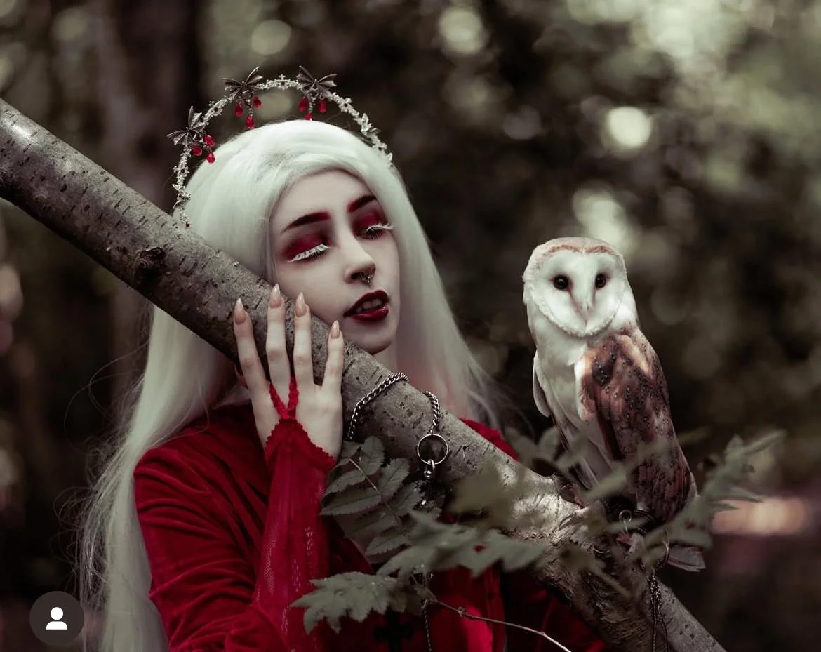 A woman with long platinum blonde hair and gothic makeup, wearing a red dress and a decorative headpiece, holding a tree branch in a dark forest with an owl perched on the branch.