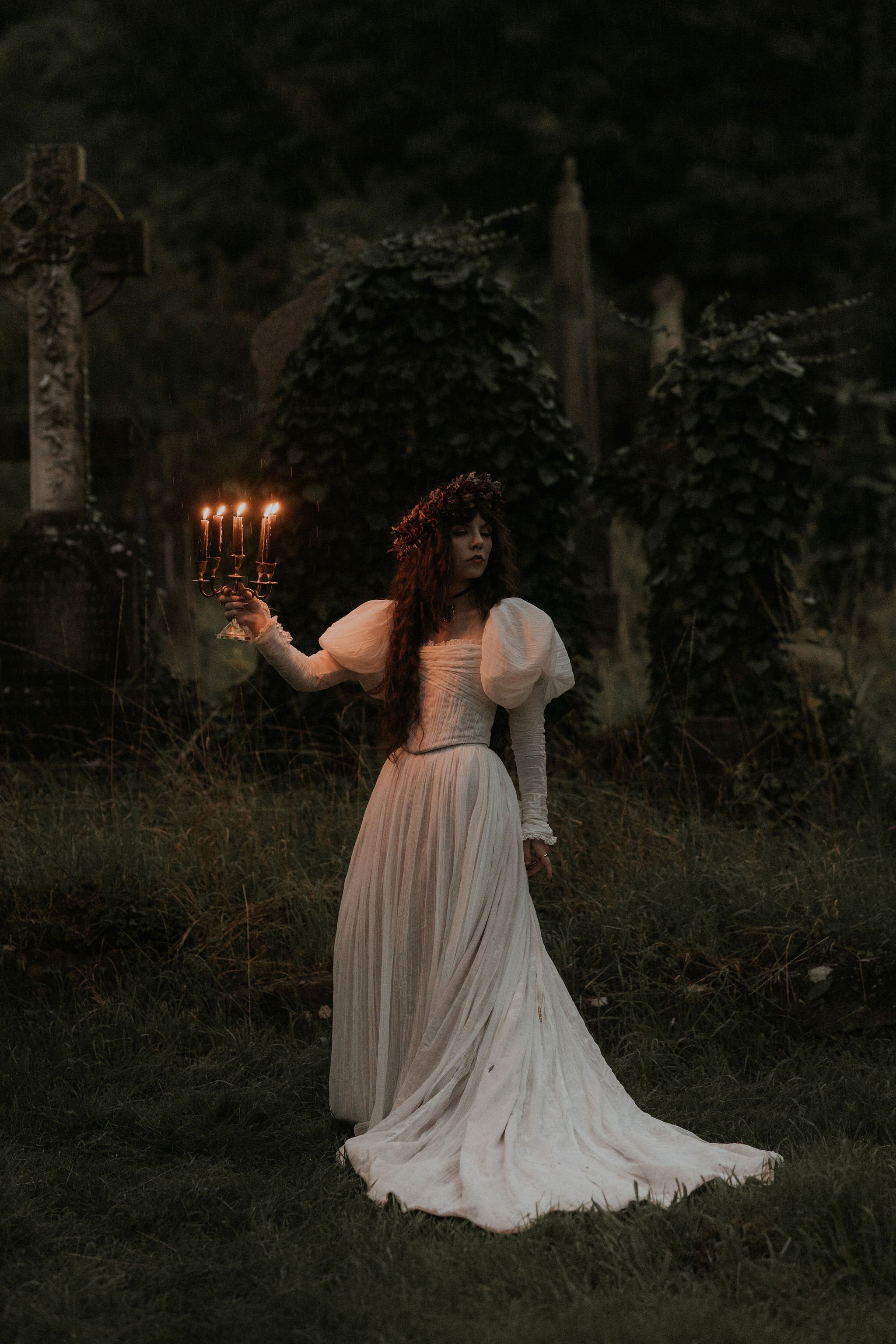 A woman in an old-fashioned white dress holding a candelabrum with five lit candles in a dark, eerie outdoor setting with an ornate cross and trees in the background.
