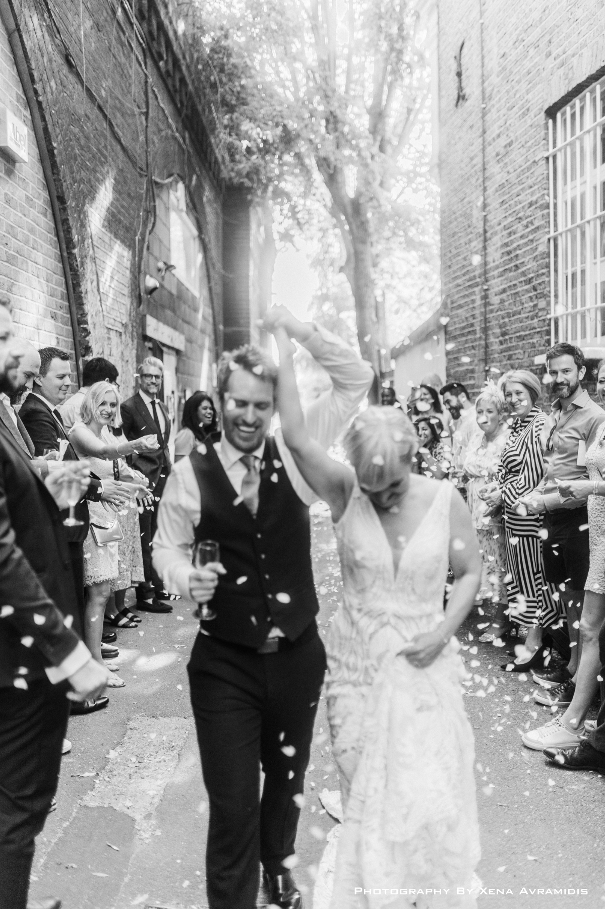 A black-and-white photo of a bride and groom celebrating their wedding while walking through a narrow alleyway, surrounded by friends and family, with confetti falling around them.
