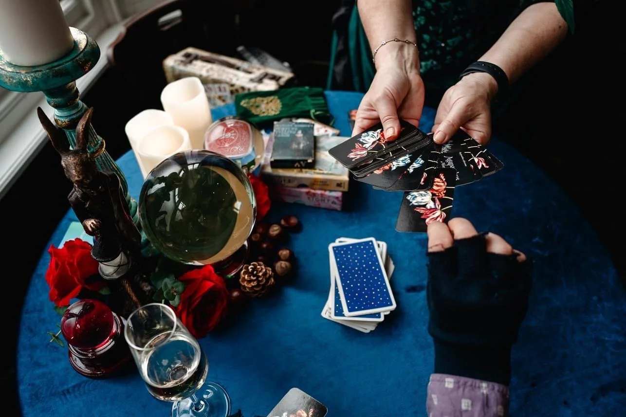 Two people playing a card game at a table covered with a blue cloth. The table has candles, a crystal ball, a small statue, flowers, and various items including tarot decks and tarot cards. One person's hands are visible holding a fan of tarot cards,