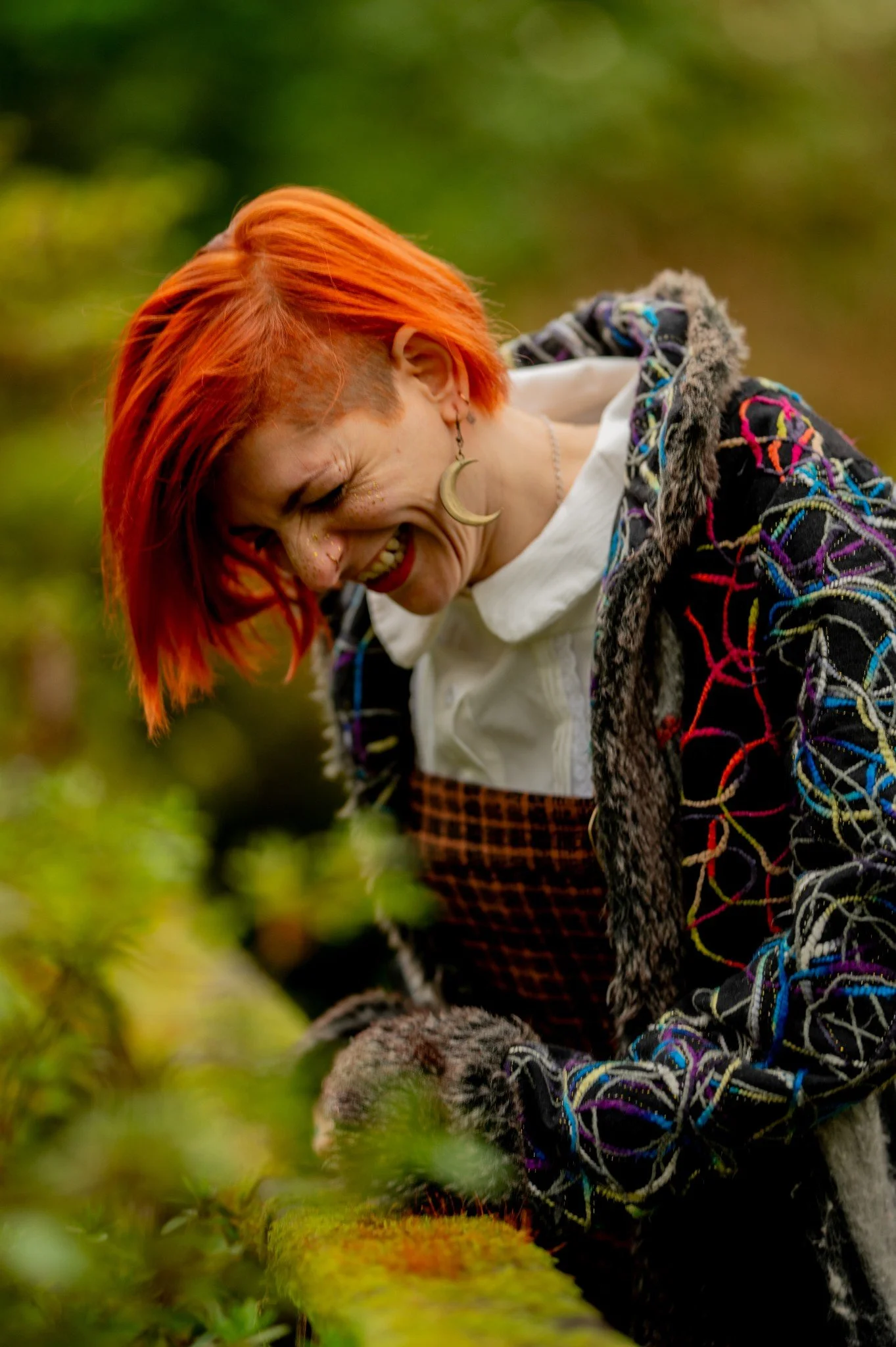 A person with bright orange hair, smiling with gold glitter on face, wearing a moon-shaped earring, colorful patterned jacket, white shirt, and checkered pants, outdoors in a natural, green setting.