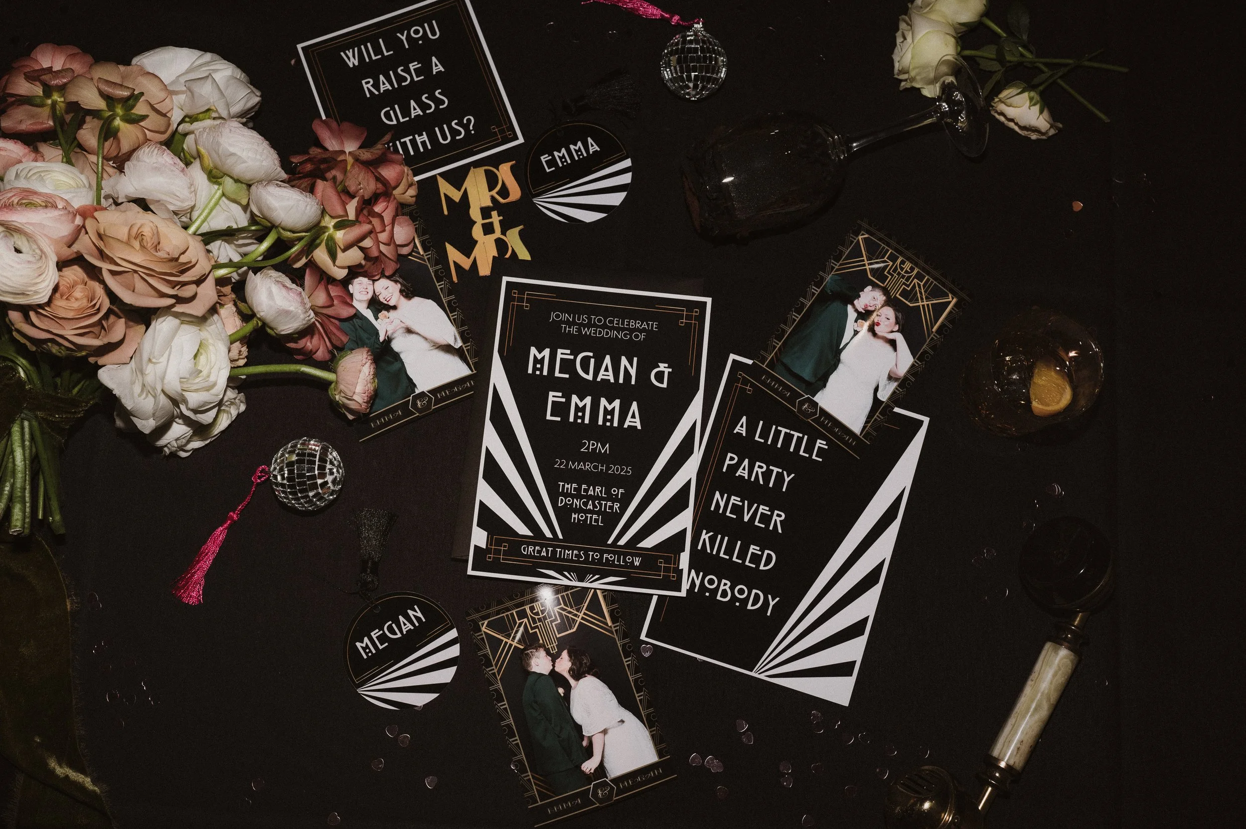 Wedding invitation and photo booth props on a black table, including a large invitation for Megan and Emma's wedding, roses, a small disco ball, vintage glasses, and a retro lipstick glamour tube.