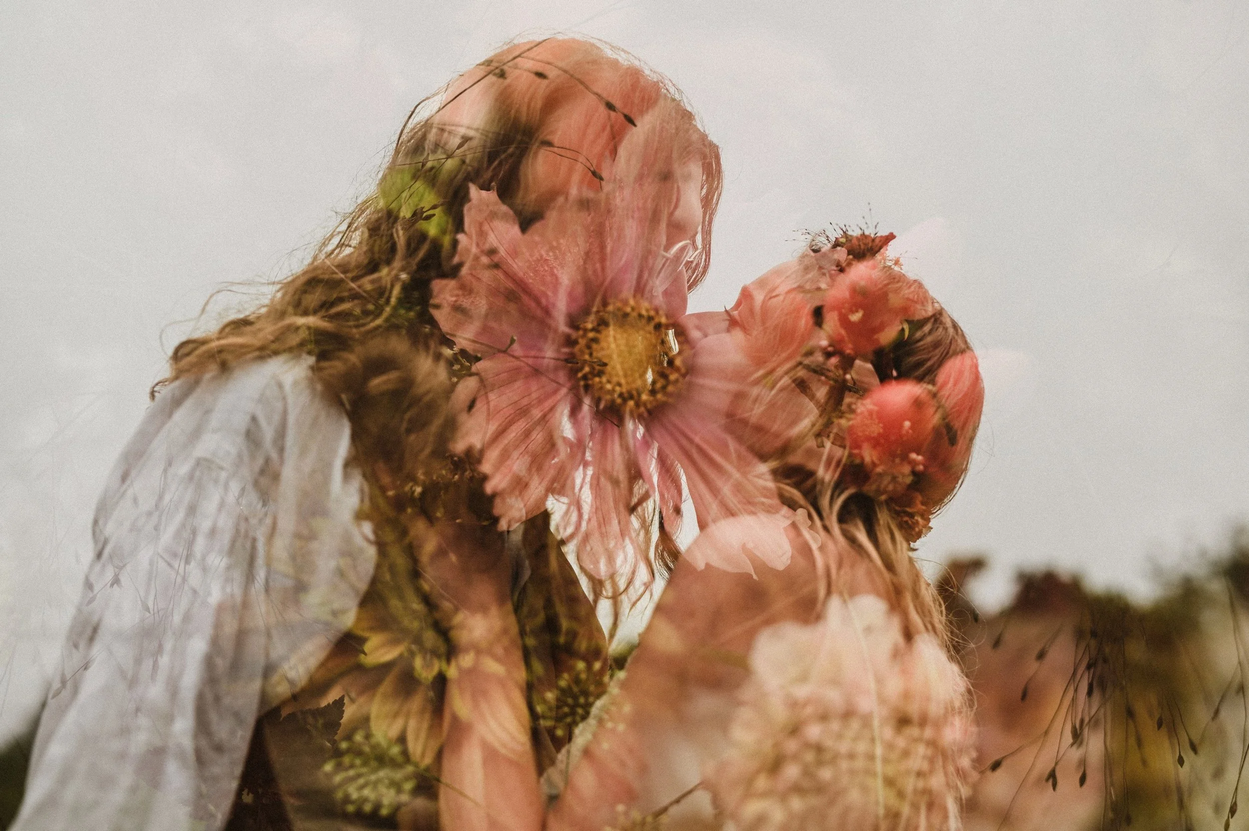 Two women with flower crowns sharing a kiss outdoors, with a double exposure effect showcasing flowers and greenery.