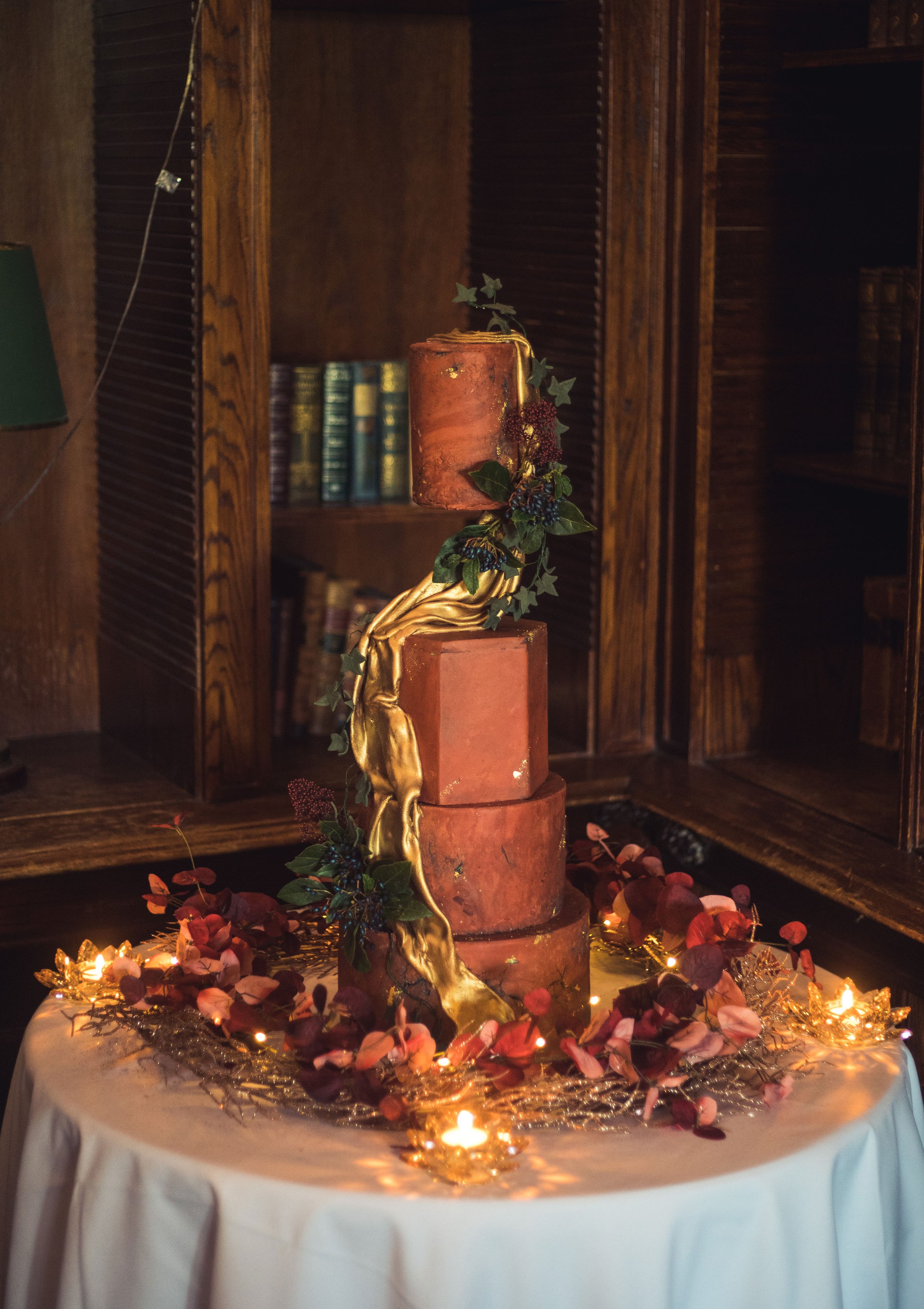 A four-tiered cake with a rustic design, decorated with gold accents and greenery, surrounded by pink and red leaves and fairy lights on a round table with a white tablecloth, set in a wood-paneled room.