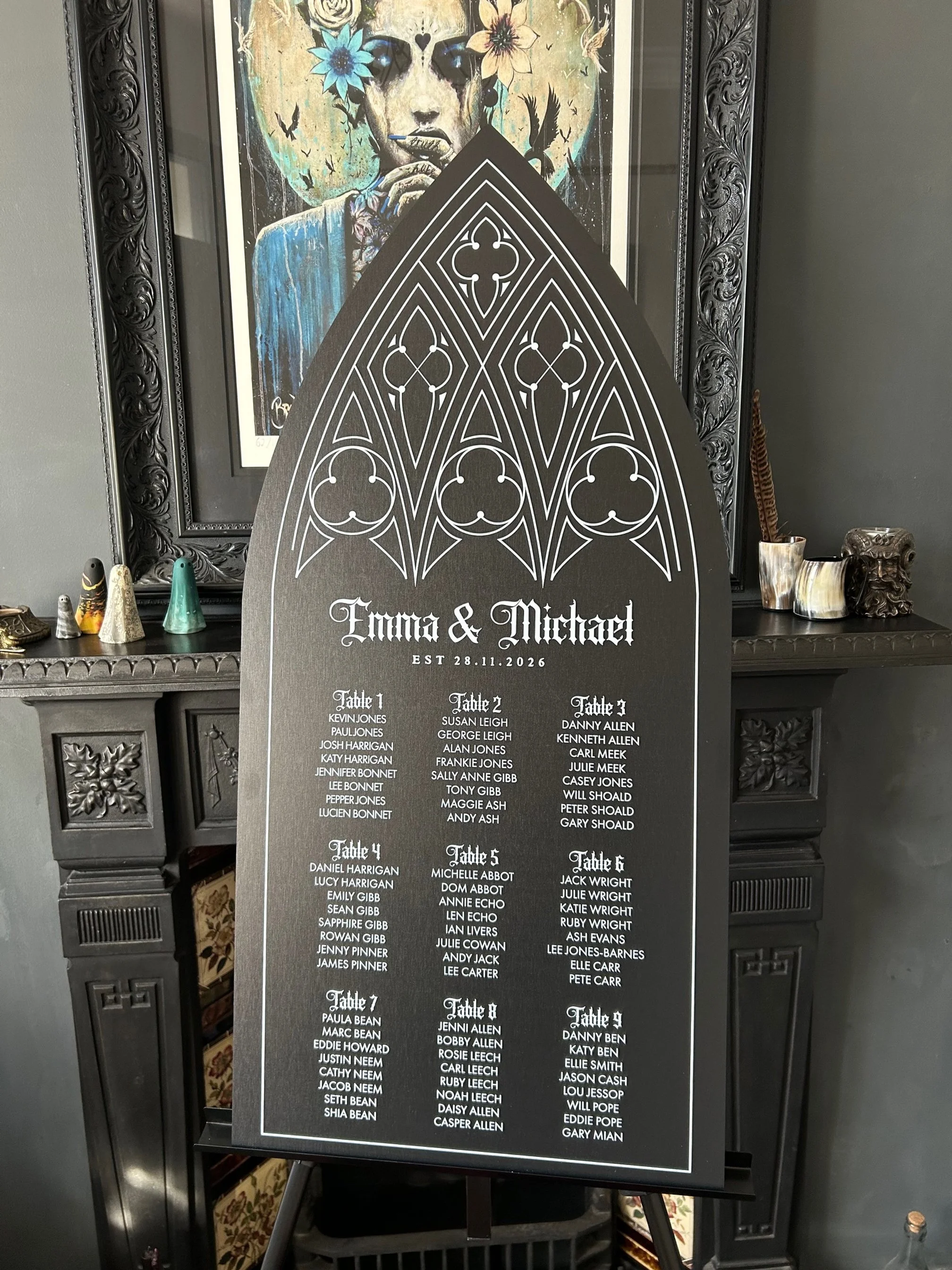 Wedding seating chart with gothic design, titled "Emma & Michael," dated November 28, 2026, showing tables with guest names, placed in front of a fireplace with decorative items and artwork.