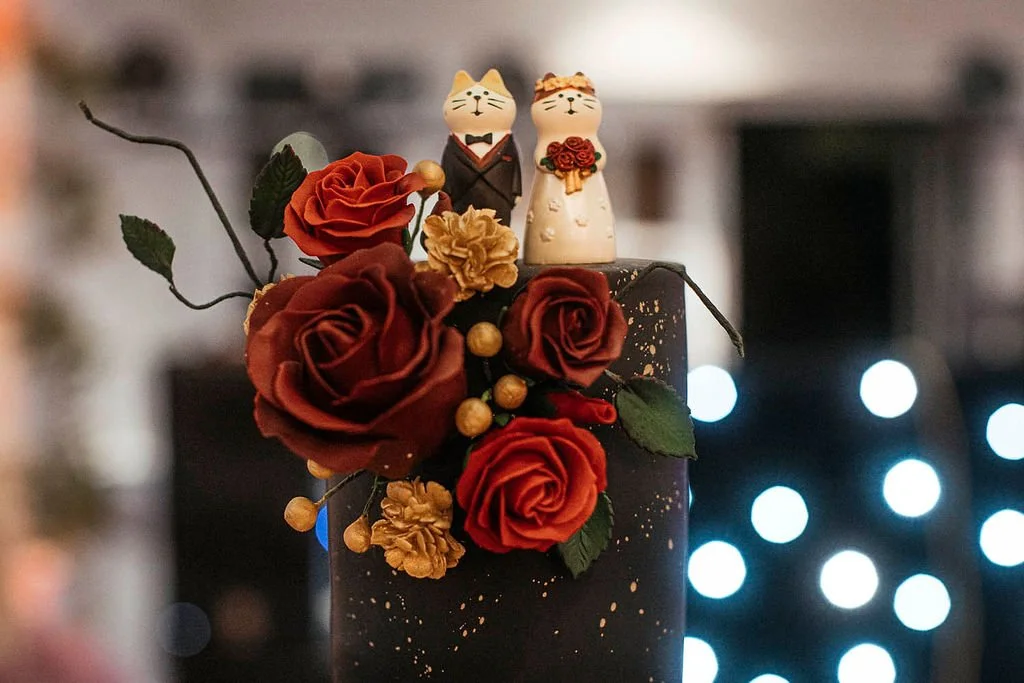 Wedding cake decorated with red and gold roses, gold berries, and two cat figurines dressed as a bride and groom.