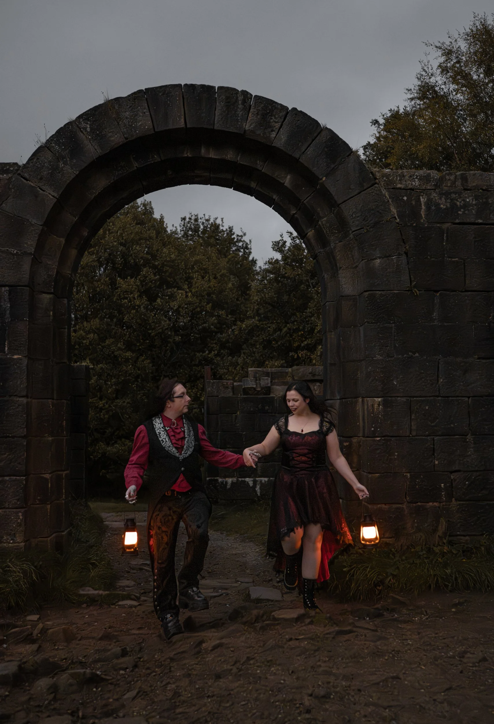 A man and a woman holding hands and dancing under a stone archway at dusk, with lanterns in their hands and trees in the background.