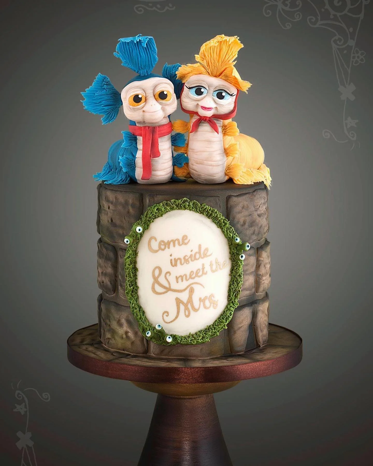 A cake designed to resemble a brick wall with two puppet-like characters on top, one with blue hair and the other with yellow hair, both with large eyes and red scarves. The front of the cake has a white oval message that reads, 'Come inside & meet the Mrs,' decorated with a green leafy border with small eyeball details.