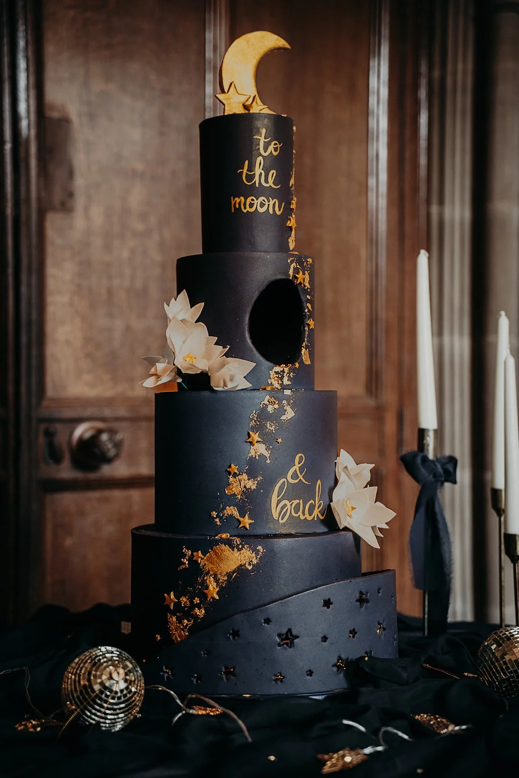 A black, four-tiered birthday cake with gold accents, decorated with white paper flowers, star-shaped confetti, and a moon and star topper. The cake features gold writing that says 'to the moon & back' and is placed on a black cloth with candles and 
