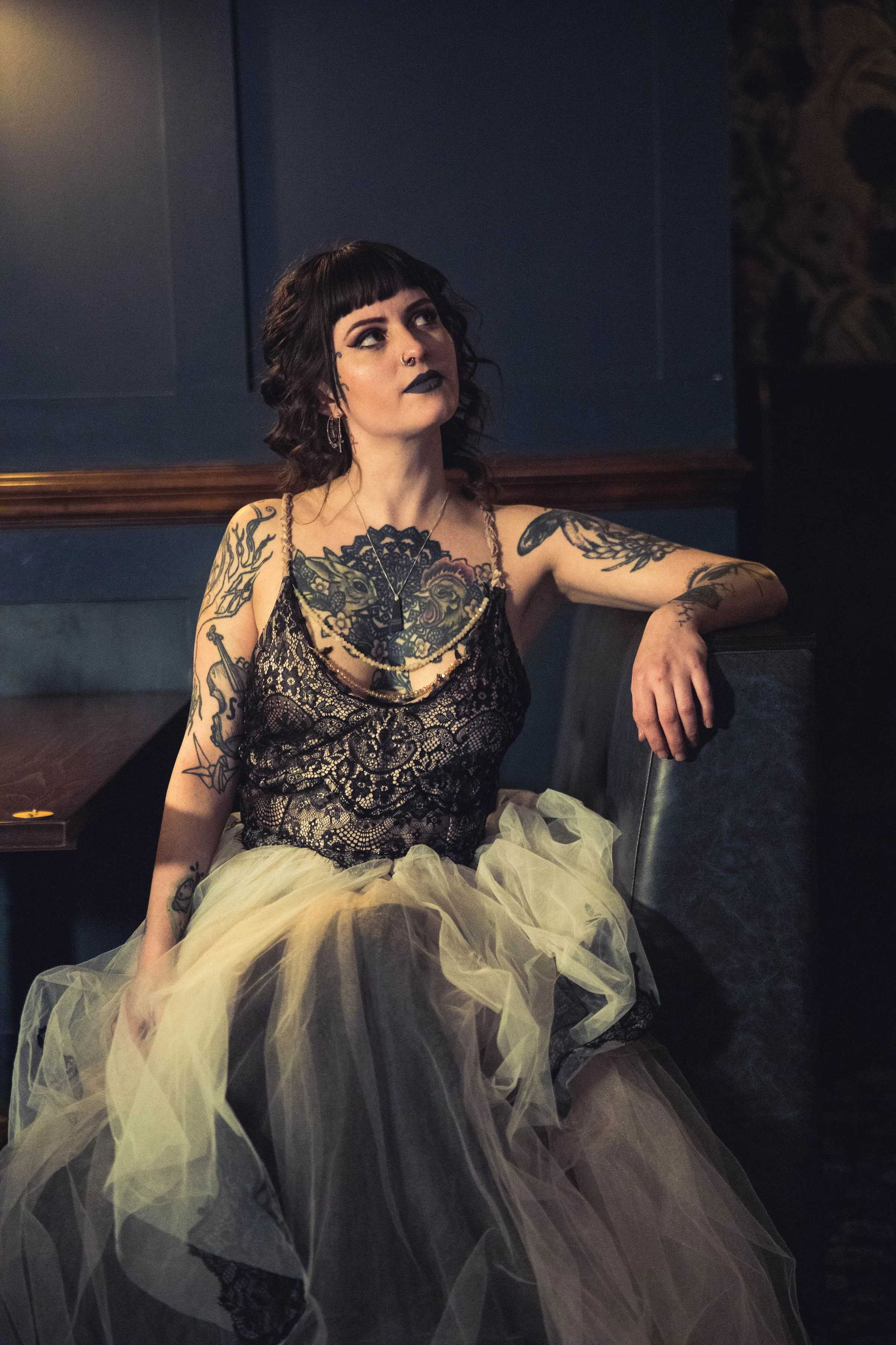 A woman with dark curly hair, tattoos, and goth makeup sitting on a booth in a dimly lit room, wearing a black lace top and a tulle skirt.
