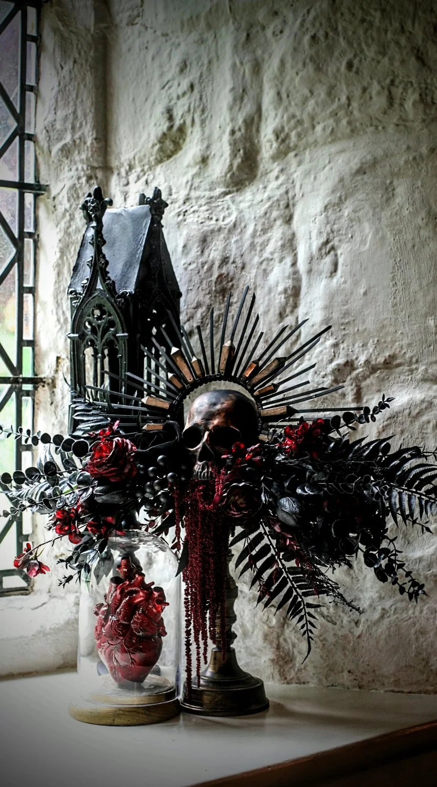 Decorative altar featuring a skull, surrounded by dark flowers, red beads, and metal rays, with a black Gothic-style structure, set against a textured stone wall with a window.