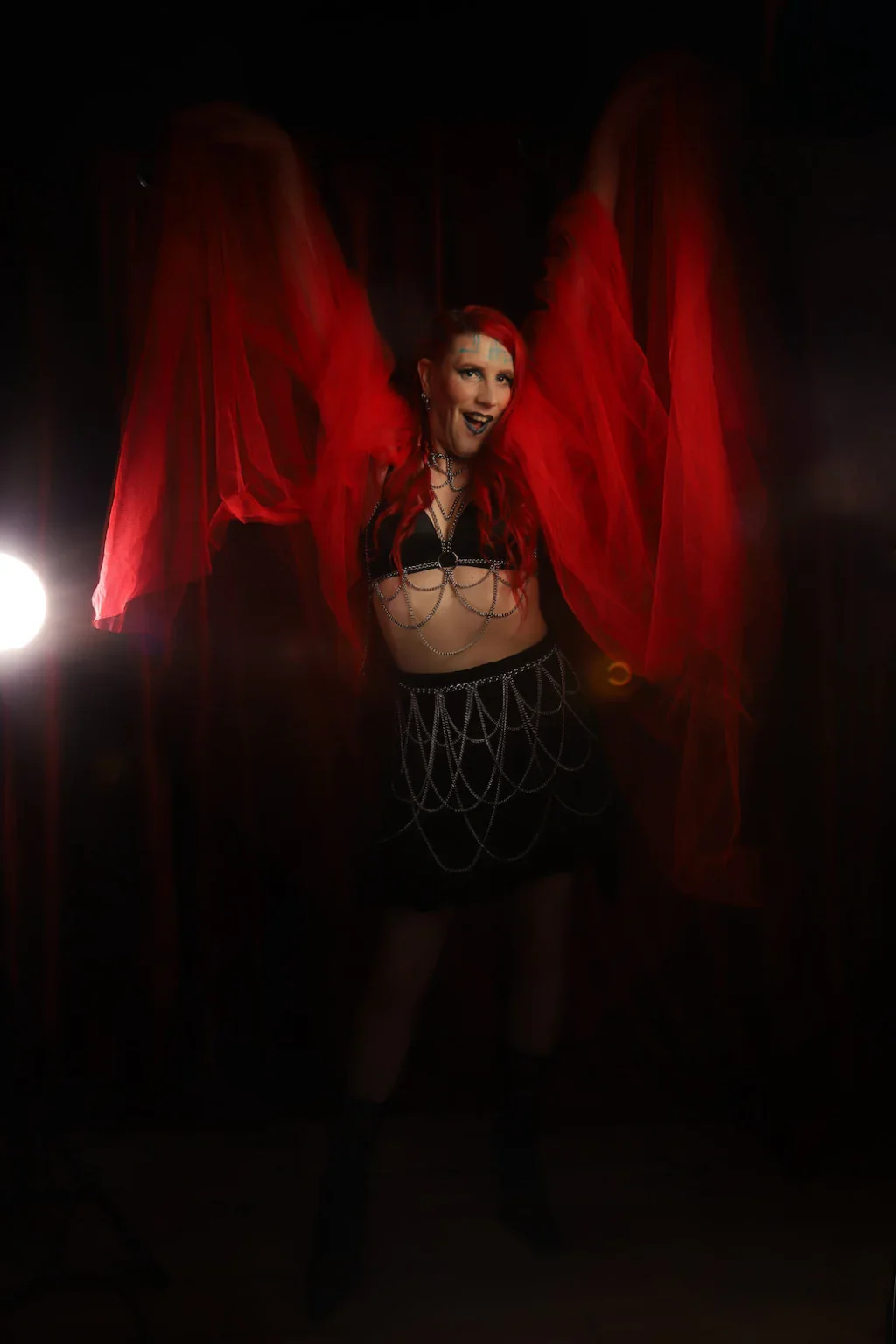 A woman in a black outfit with chains, red hair, and dark lipstick, posing with red fabric wings or drapes in a dark setting with bright lights.