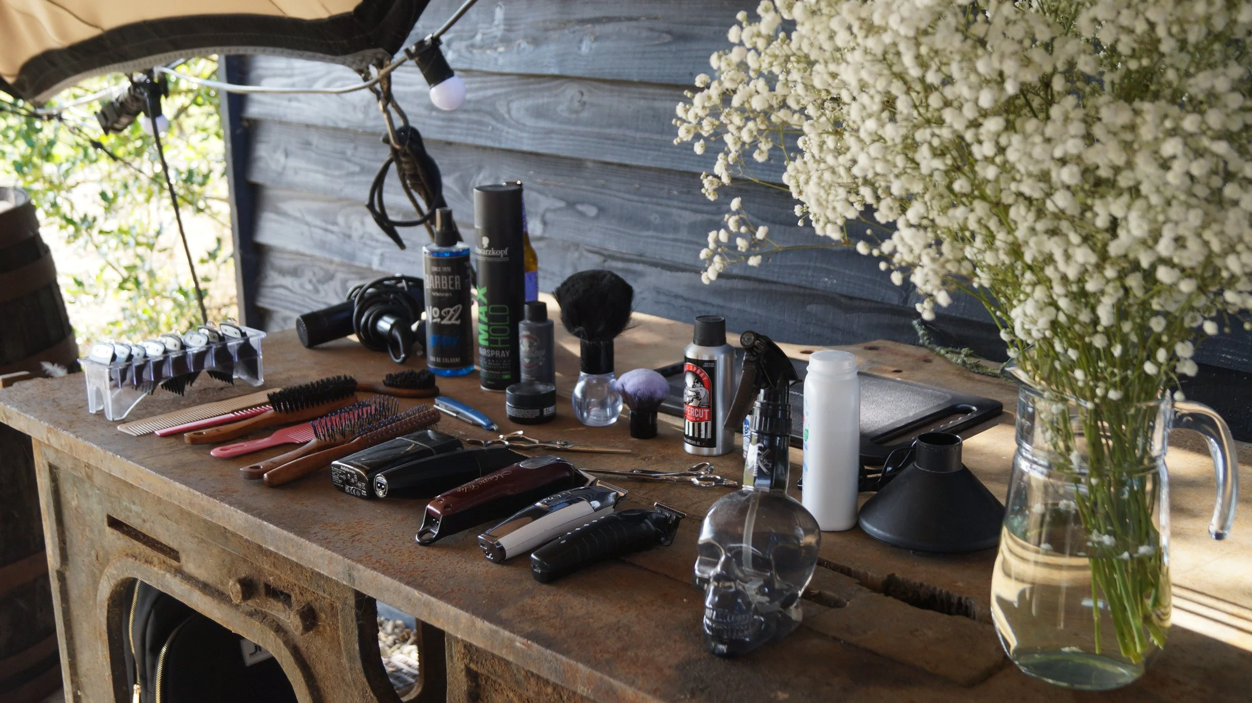 A rustic wooden table with various barber tools and products, including clippers, brushes, spray bottles, and a skull ornament, is next to a large vase of white baby's breath flowers.