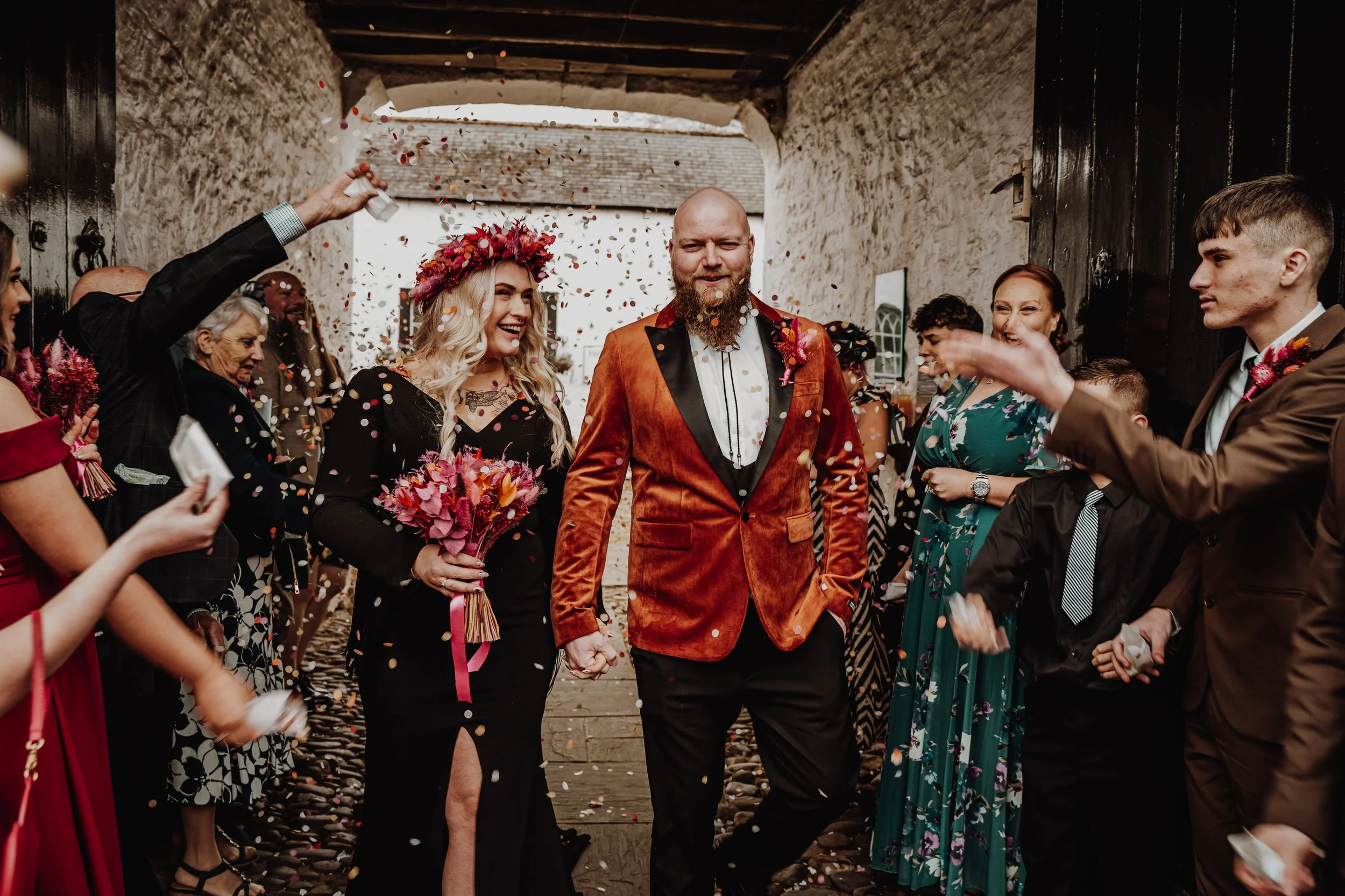 Bride and groom walking hand in hand through a shower of confetti surrounded by wedding guests in a rustic outdoor setting