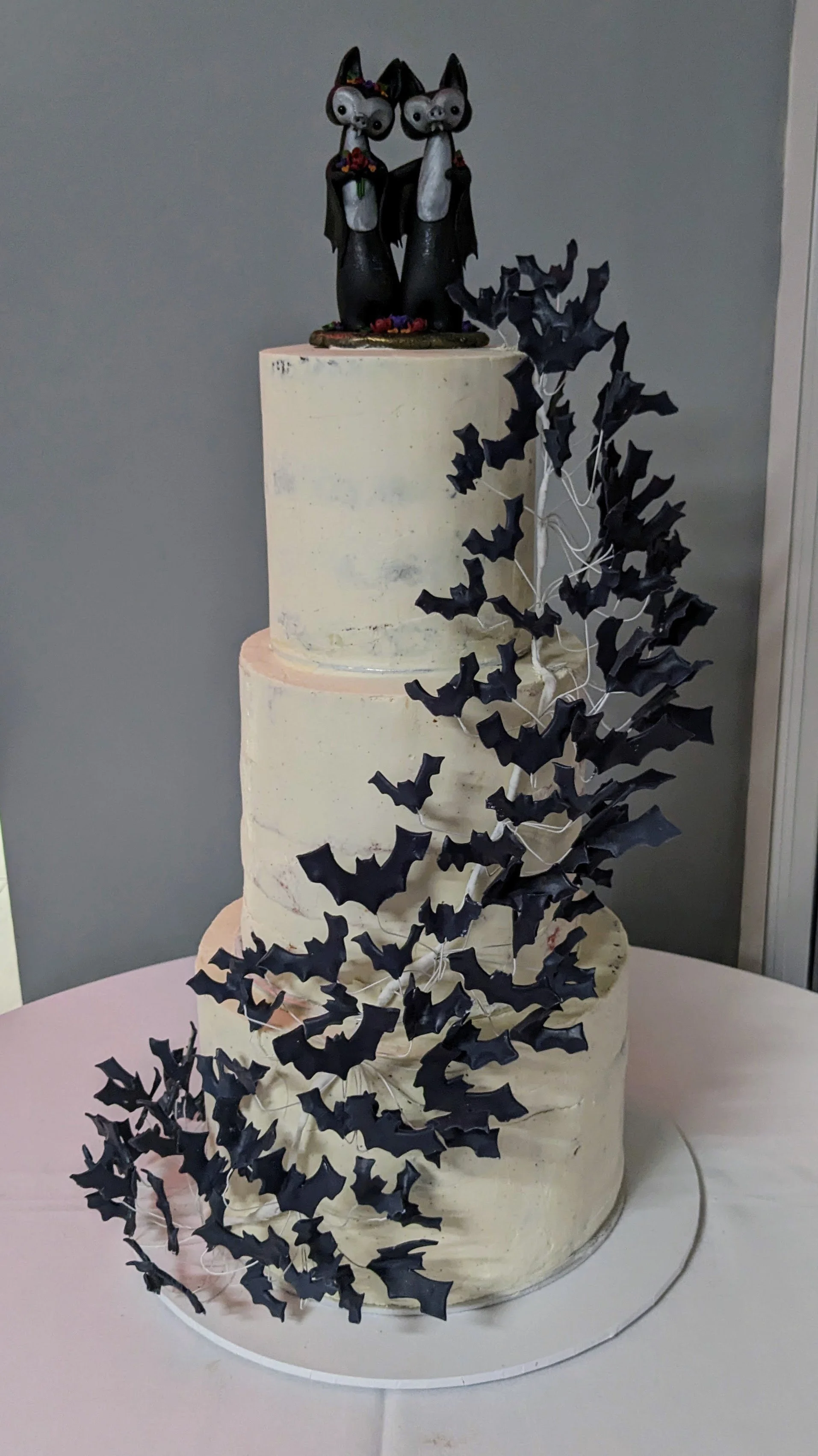 A three-tiered white cake decorated with black paper bats and topped with two black cat figurines dressed as witches, with bats arranged around the cake.