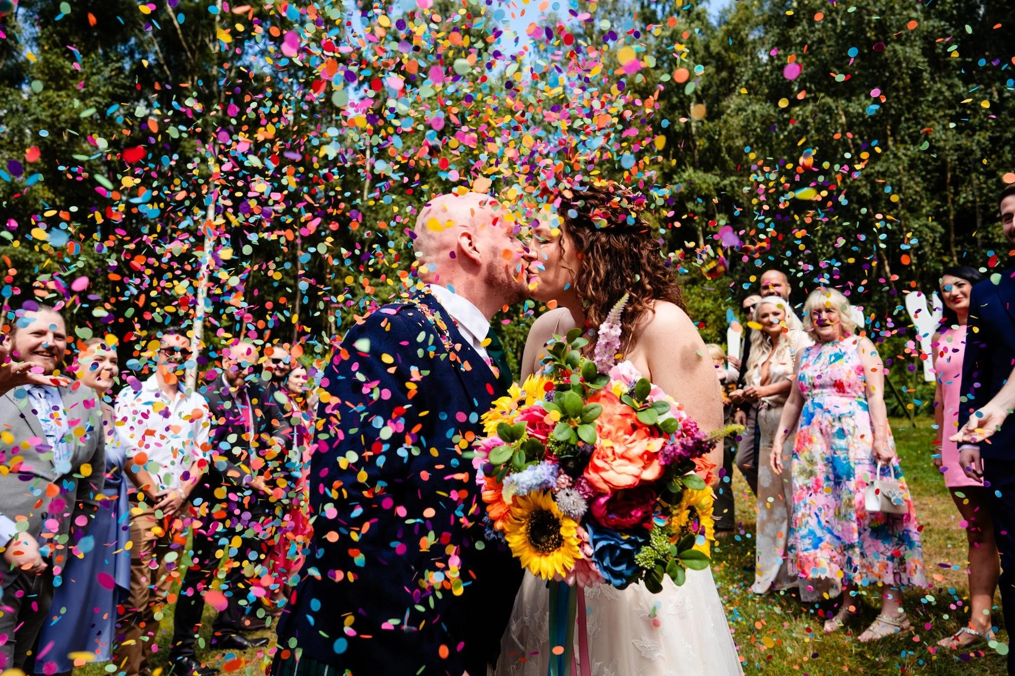 A couple kissing at their outdoor wedding, surrounded by friends and family, with colorful confetti falling around them.